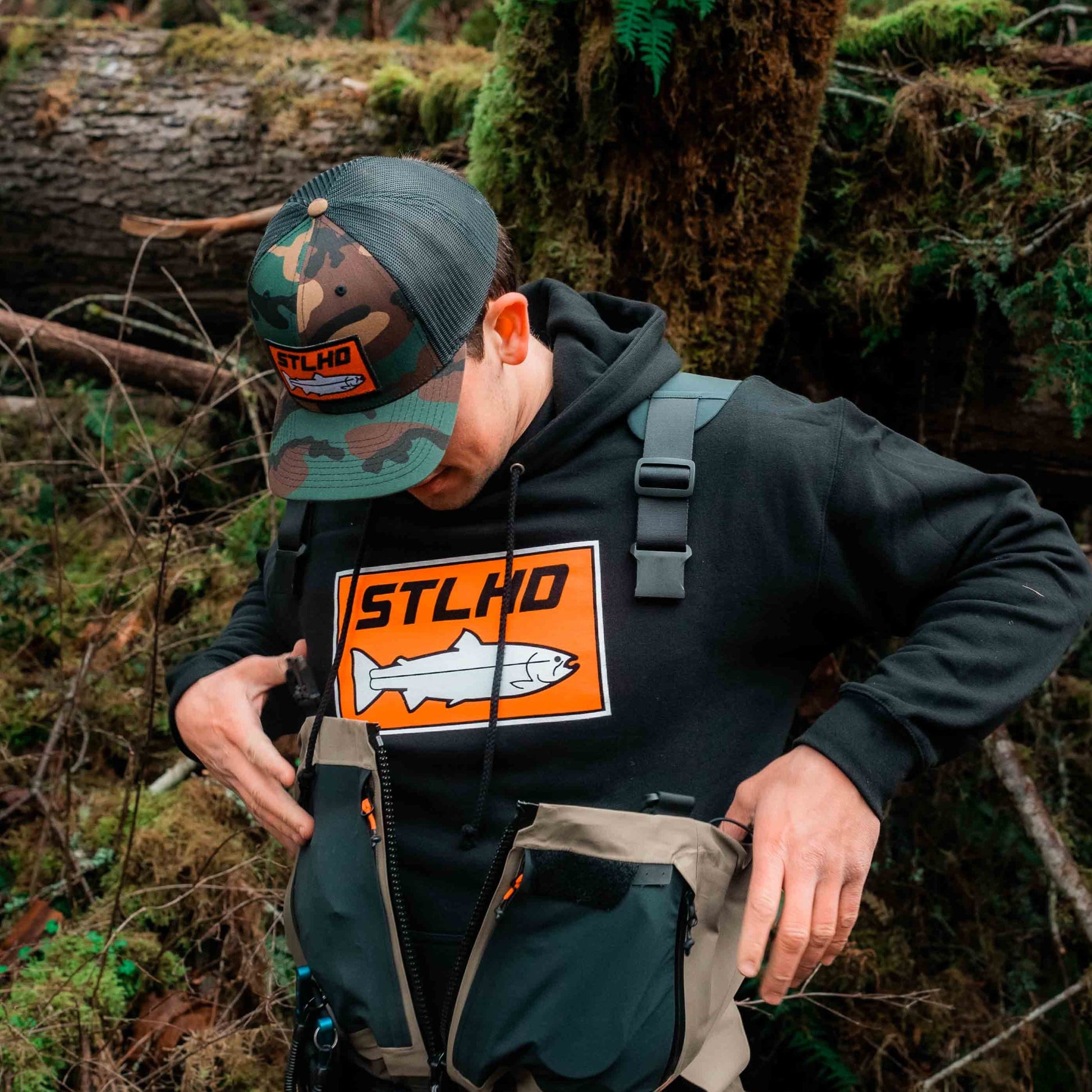 A person in a forest adjusts their tan fishing vest while wearing an STLHD GEAR Mens Standard Logo Black Premium Hoodie and a camo STLHD hat. Mossy logs and green foliage are visible in the background.