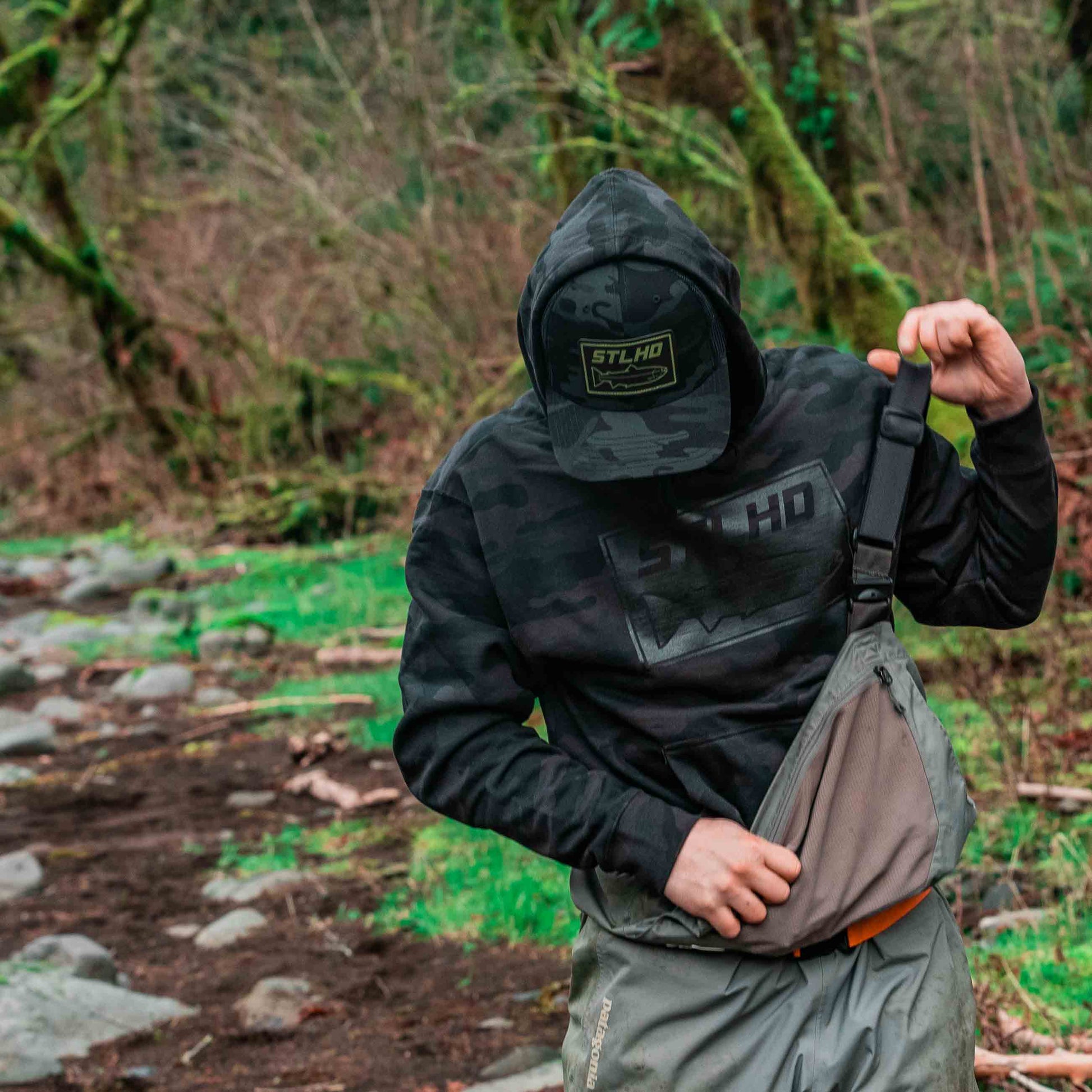 A person wearing the STLHD GEAR Black Water Black Camo Standard Hoodie stands in a wooded area, adjusting the strap of gray overalls near a rocky path. Their face is hidden by the hood and cap.