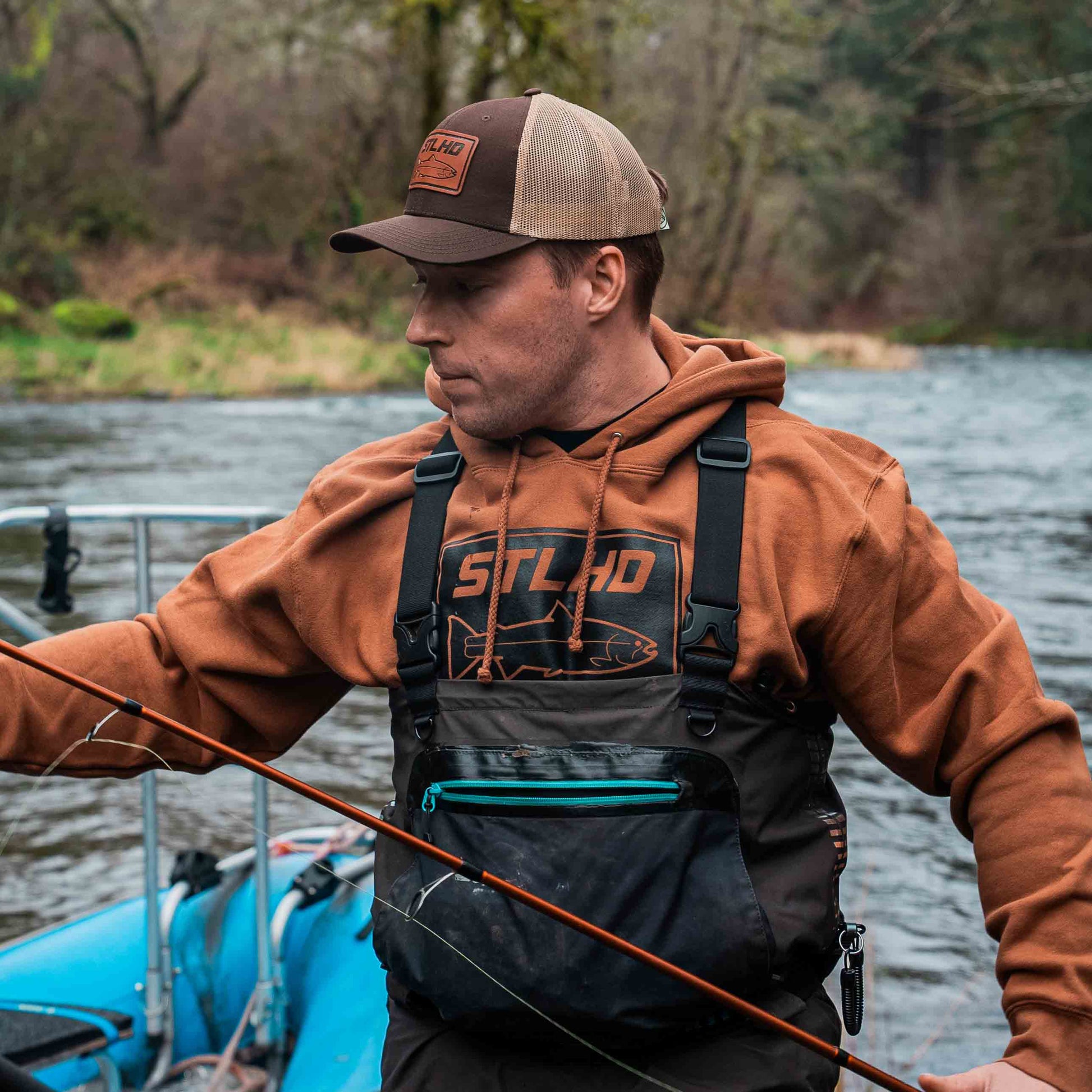 Wearing the STLHD GEAR Mens Yuba River Saddle Brown Premium Hoodie, a man stands on a boat with fishing waders and rod, enjoying the river scenery. The ultra-soft fleece lining keeps him comfortable all day outdoors.