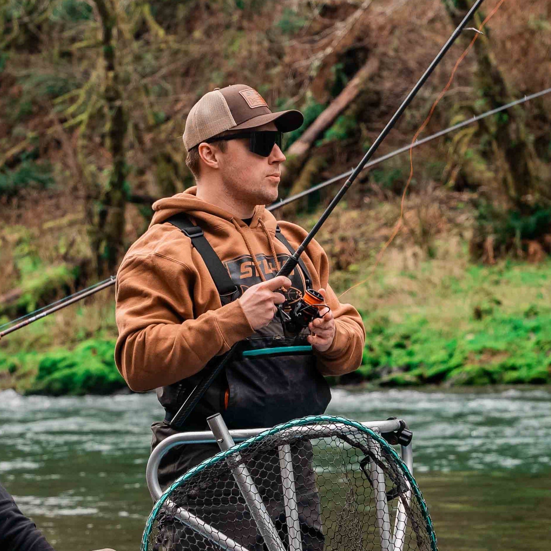 A man in an STLHD GEAR Mens Yuba River Saddle Brown Premium Hoodie stands by a river with a fishing rod and net, surrounded by trees and greenery, wearing a cap and sunglasses.
