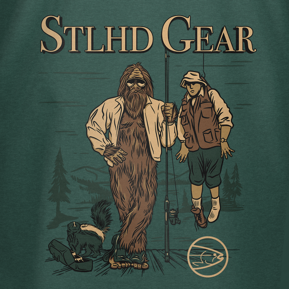 An illustration of Bigfoot with a fishing rod stands by a fisherman, a skunk nearby and pine trees behind. “STLHD GEAR” is above—featured on the STLHD Men’s Lake Potowotominimac T-Shirt for breathable comfort.