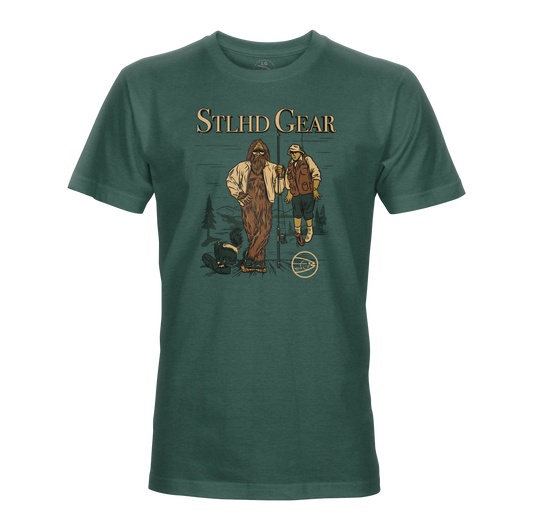 The STLHD Men’s Lake Potowotominimac T-Shirt by STLHD GEAR features the brand logo above Bigfoot and a fisherman in the woods, made with breathable CHROMELITE fabric. A small STLHD logo appears near their feet.