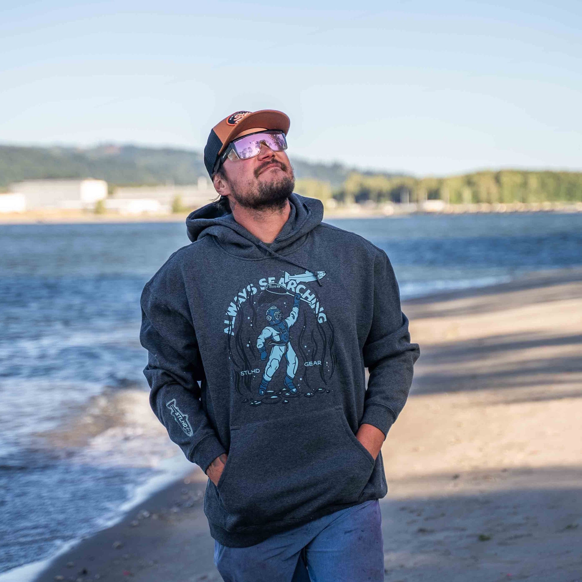 A man in sunglasses, a cap, and an STLHD GEAR STLHD Men’s Deep Dive Premium Hoodie featuring an astronaut graphic walks along a sandy beach near the water, glancing up at the sky with trees and buildings in the background.