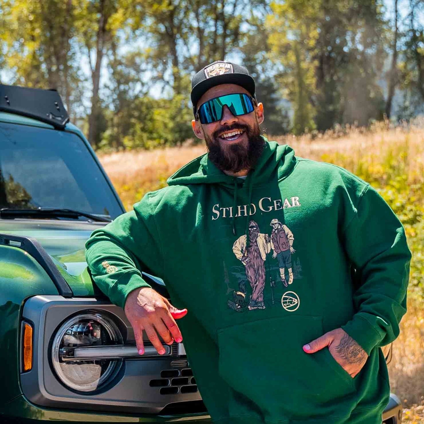 Man wearing a green hoodie with Bigfoot holding up a man with a fishing rod, with STLHD GEAR TEXT across the top, standing next to a vehicle in a natural setting.