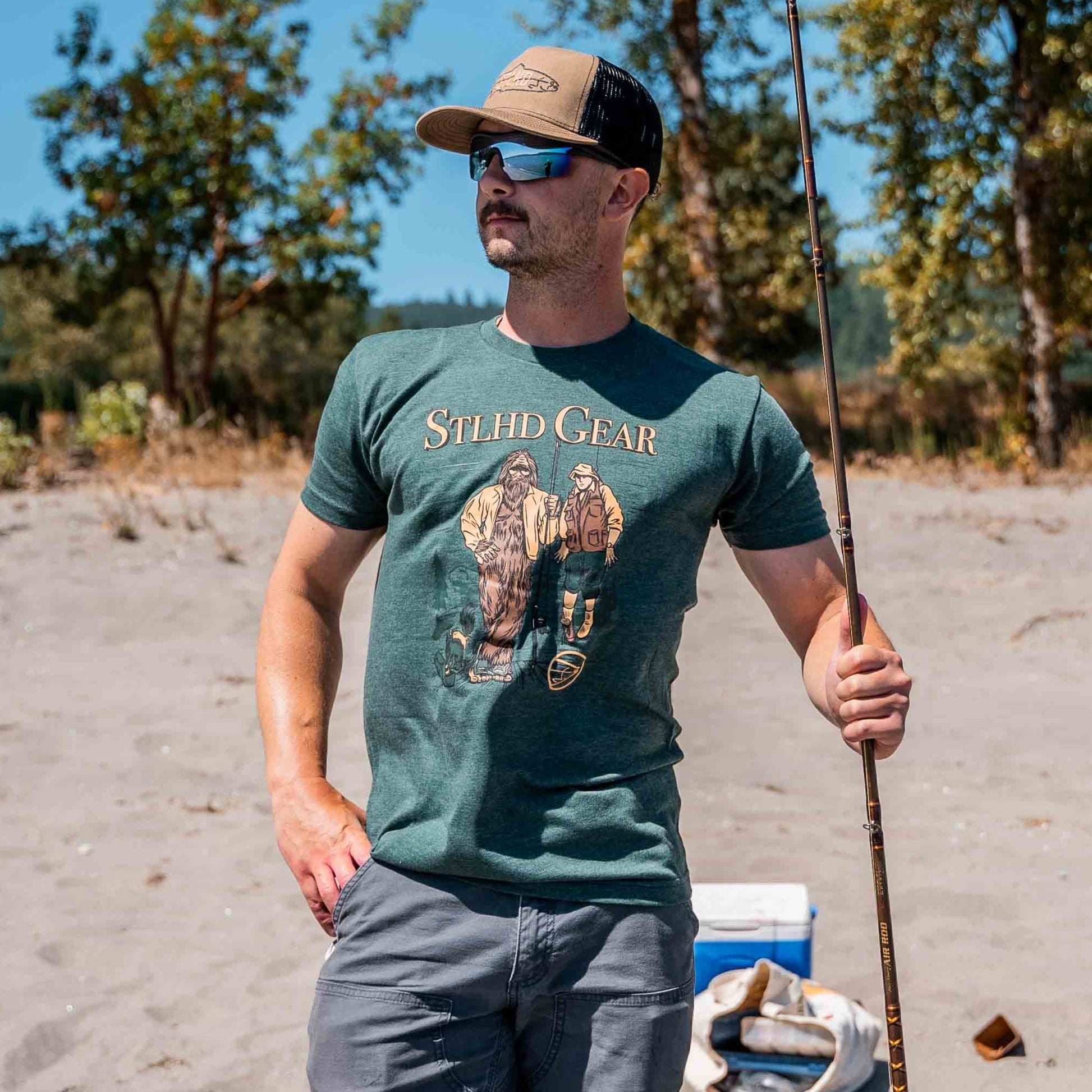 A man in sunglasses and a cap wears the STLHD GEAR Men’s Lake Potowotominimac T-Shirt, holding a fishing rod on a sandy beach with trees and a cooler in the background, enjoying breathable comfort.