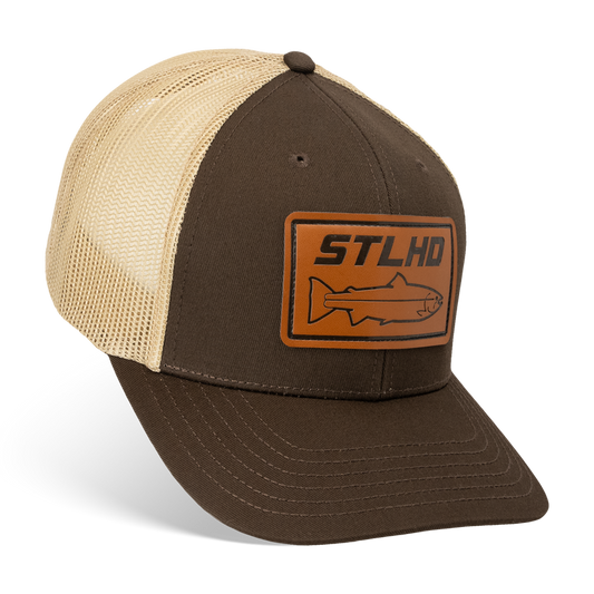 The STLHD GEAR Steelhide Brown/Khaki Hat is a mesh trucker hat with a brown and beige design, a rectangular leather patch displaying STLHD and a fish outline on the front, plus an adjustable snapback for a custom fit.