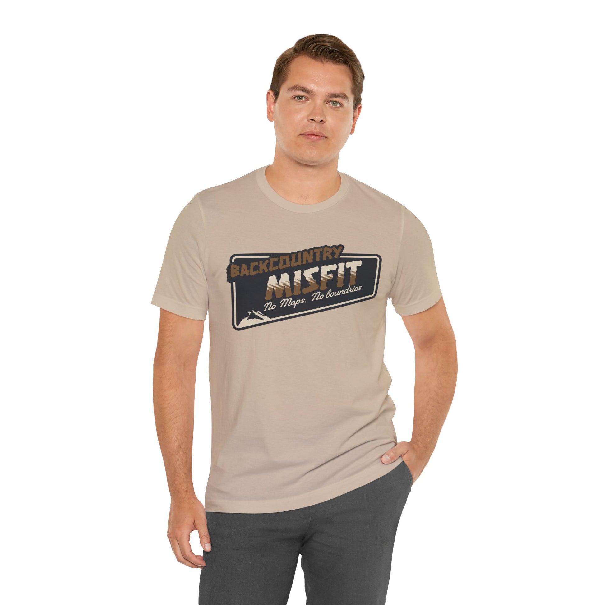 A man with short brown hair models the Printify Backcountry Misfit T-Shirt, a rugged beige hiking tee for trail rebels and off-grid wanderers. The shirt reads No Maps. No Boundaries. He stands with one hand in his pocket, looking at the camera.