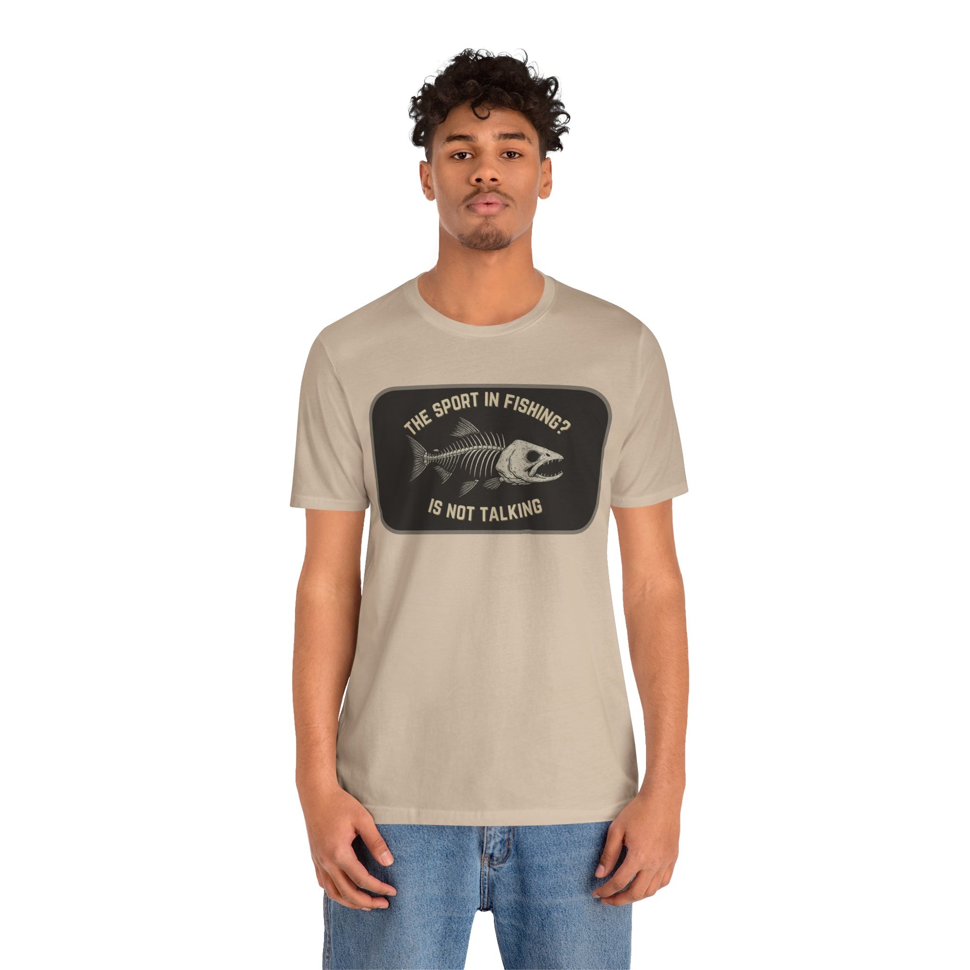 A young man with short curly hair models Printify’s “The Sport in Fishing?” Tee, a beige unisex shirt with a distressed fish skeleton print and slogan, against a plain white background as he looks at the camera.