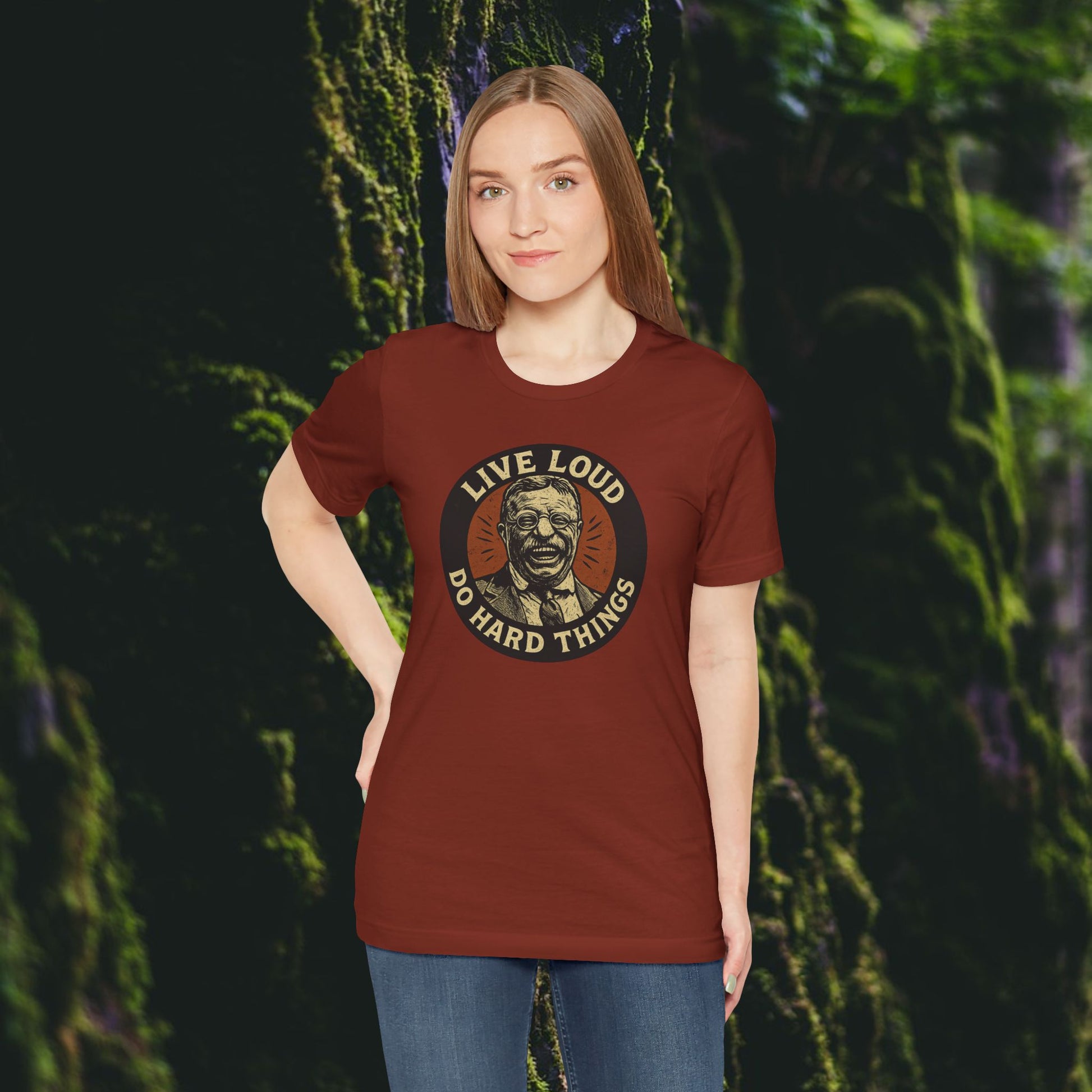 A woman stands before a mossy forest, wearing the Printify Live Loud, Do Hard Things Teddy Roosevelt Tee in maroon, featuring an illustrated face and bold lettering.