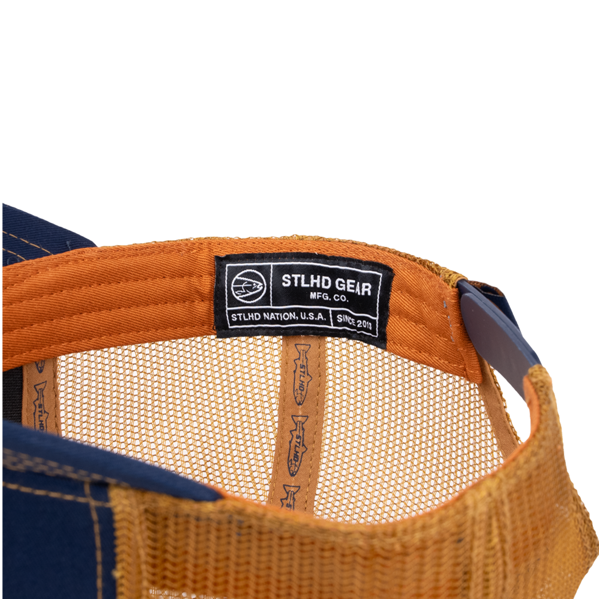 Close-up of the STLHD Let’er Rip Trucker Hat by STLHD GEAR, showing a black brand label stitched on an orange band, tan mesh panels, and blue fabric—perfect for outdoor adventures.