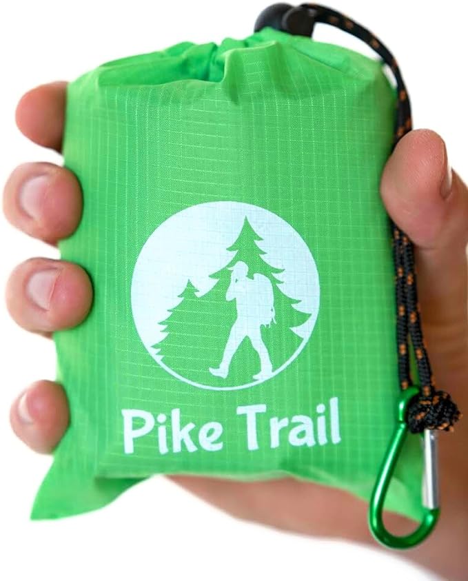 A hand holds the Pike Trail Pocket Blanket—Compact, Rugged, Always Ready—in a small green drawstring pouch with a hiker logo. A black and green carabiner is attached for easy carrying on outdoor adventures.