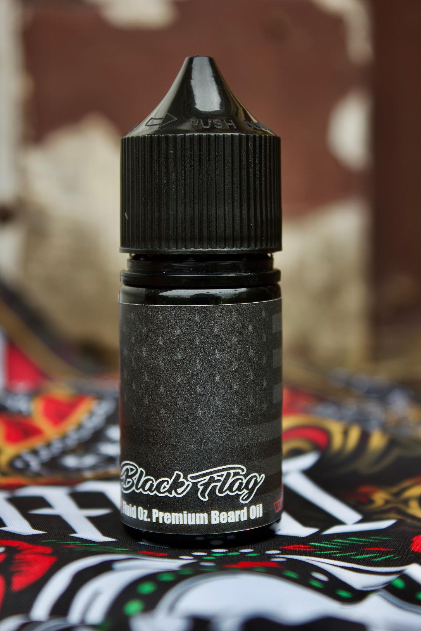 A small black bottle labeled BLACK FLAG Beard Oil & Balm by Bedlam Beard Company stands upright on a colorful patterned surface, set against a blurred brown and white background that hints at its masculine fragrance.