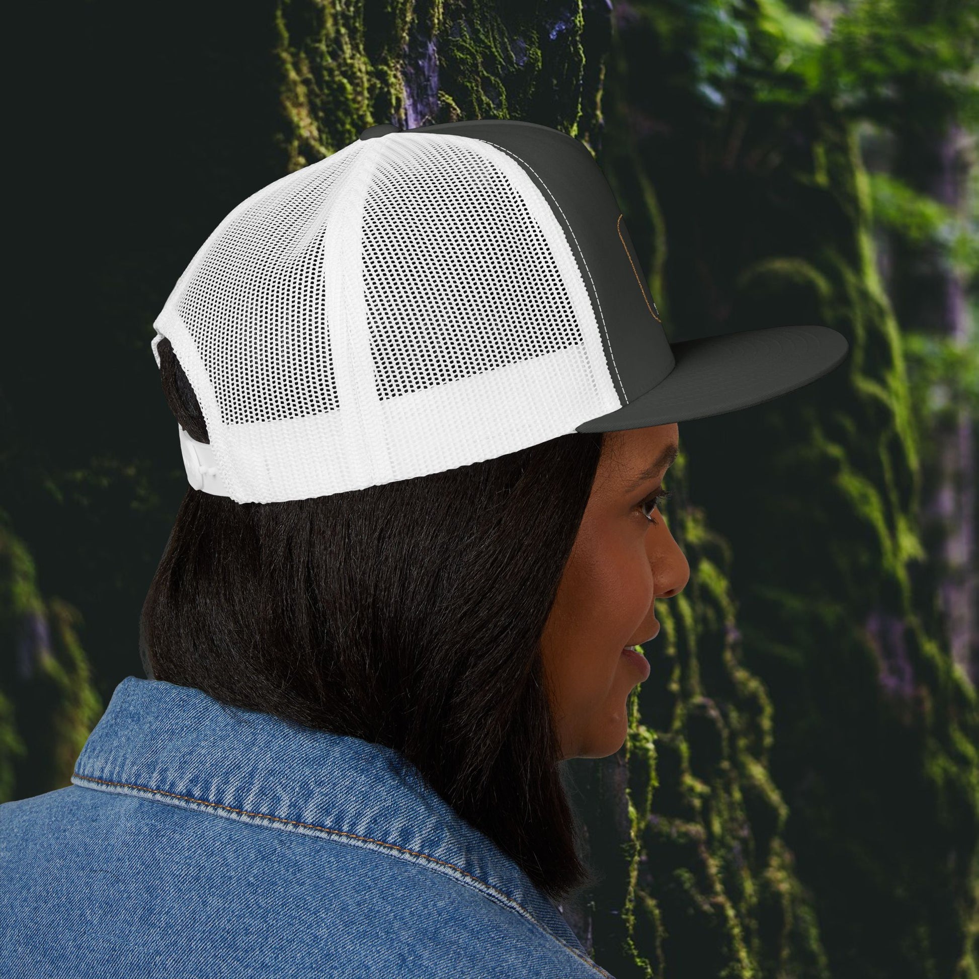 A person in profile wears a Printify Iron Pines Trucker Hat – “Live Loud. Be Inconvenient.” Brass Knuckle Edition, with white and black mesh, and a blue denim jacket, standing by green moss-covered rocks.