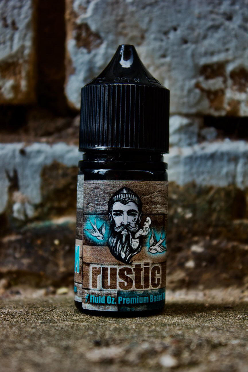 A small bottle of Bedlam Beard Company Rustic beard oil—Coffee, Tobacco Flower, Rum, Vanilla & Black Cherry—featuring an illustrated bearded man and leafy accents on the label, set against a weathered brick wall background.