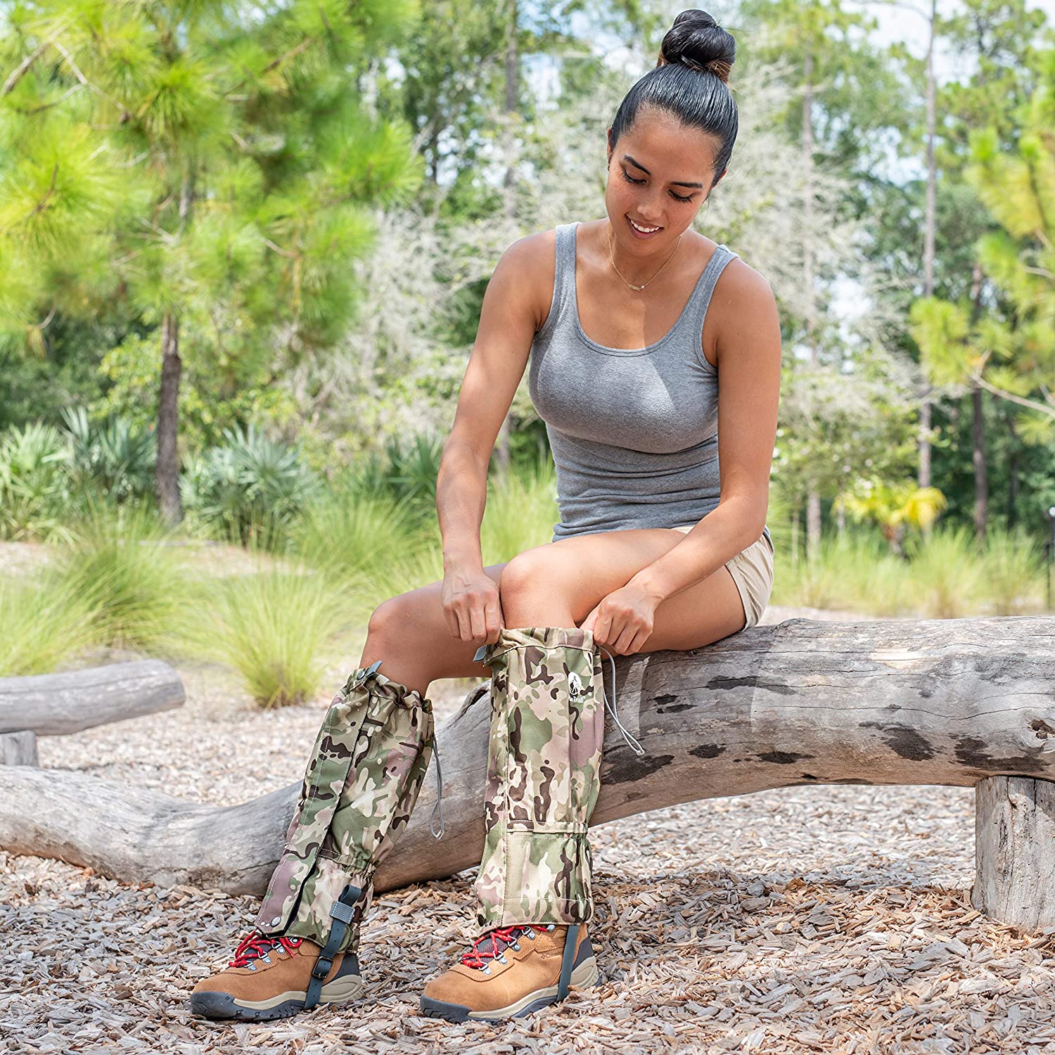 A woman in a gray tank top and shorts sits on a log outdoors, smiling as she puts on Pike Trail All-Season Waterproof Leg Gaiters—built for the trail, the hunt, and everything in between—over her hiking boots amid lush greenery.