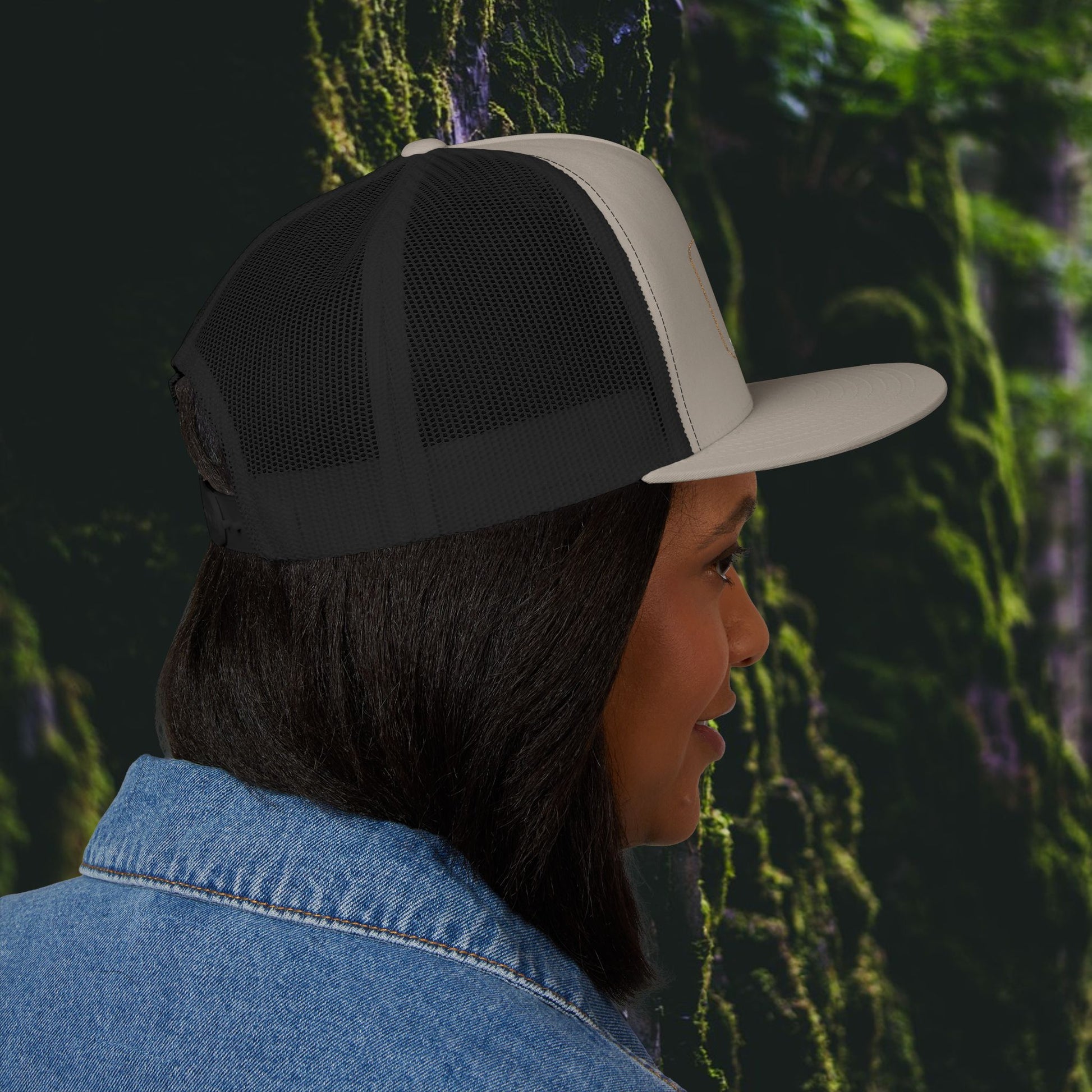 A woman with straight dark hair wears the Printify Iron Pines Trucker Hat – “Live Loud. Be Inconvenient.” Brass Knuckle Edition and a blue denim jacket, shown in profile against a lush, green forest background.