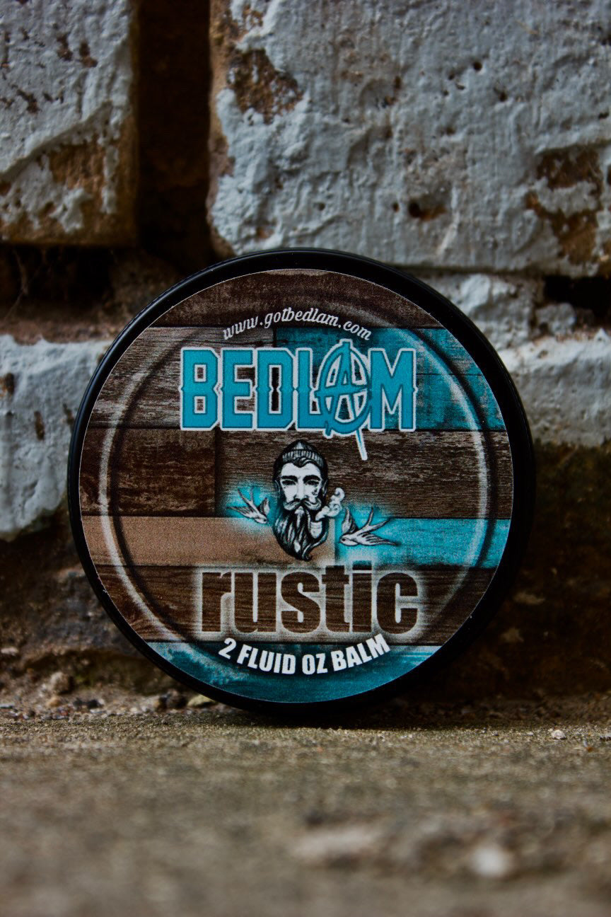 A round tin of Bedlam Beard Company’s Rustic—Coffee, Tobacco Flower, Rum, Vanilla & Black Cherry—features an illustrated bearded man on a blue and wood-toned label, set against a weathered brick wall.