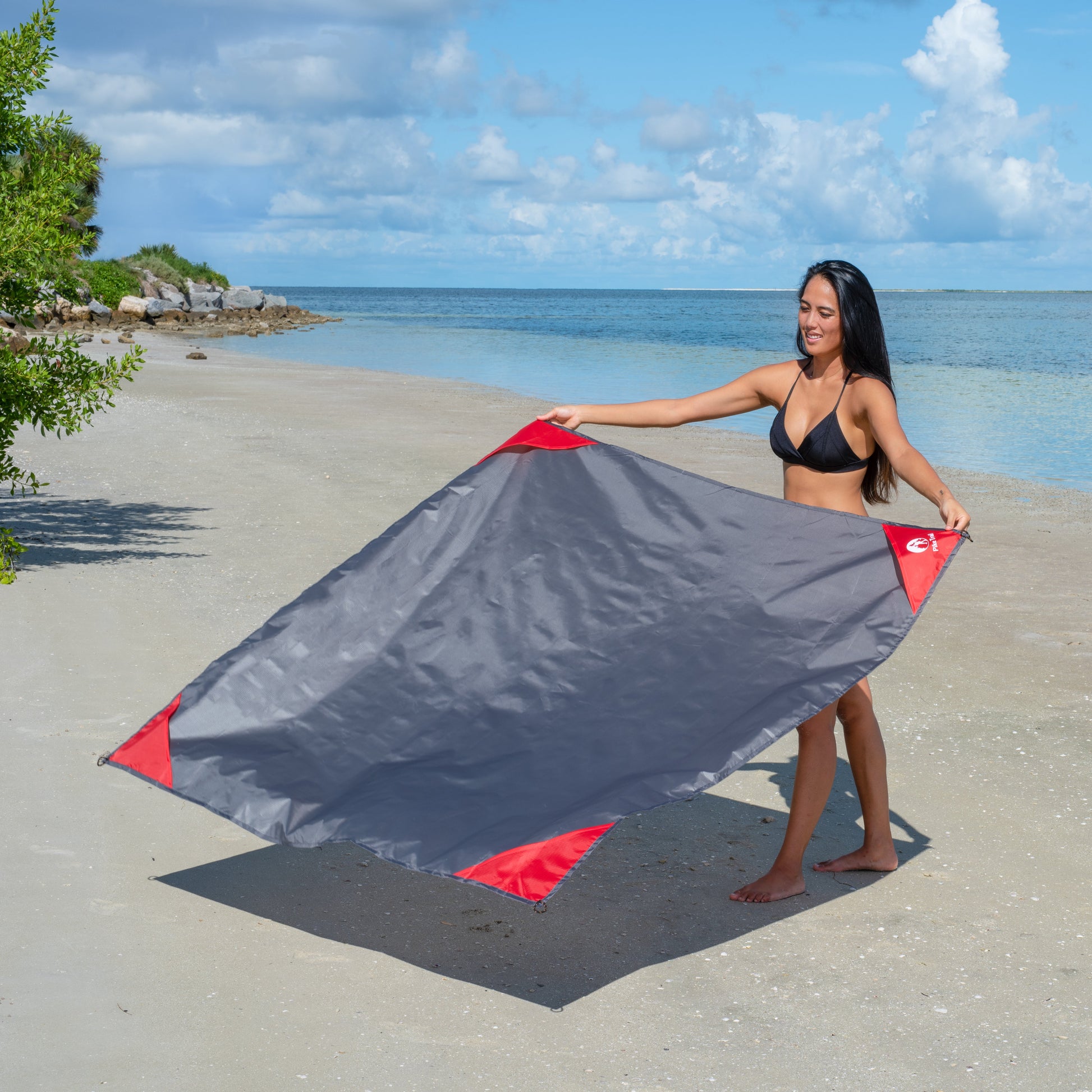 A woman on a sandy beach by the ocean unfolds the Pike Trail Pocket Blanket in gray and red. This compact, rugged blanket from Pike Trail is always ready for outdoor adventures. Partly cloudy skies and greenery complete the scene.