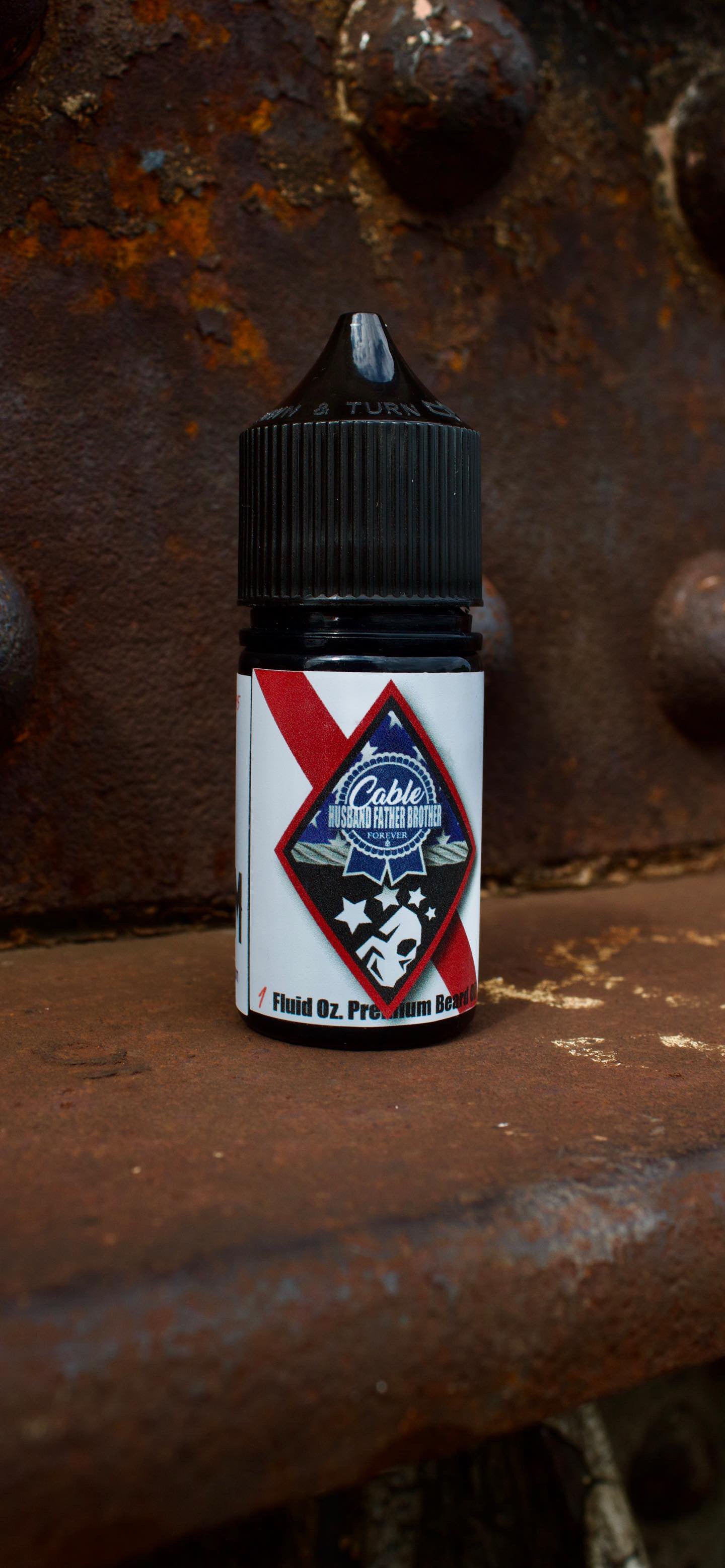 A small bottle of CABLE - Warm & Spicy Blend by Bedlam Beard Company, with a black cap and a white label featuring a blue diamond logo and red diagonal stripe, rests on rusty metal bolts, evoking a bold, masculine scent.