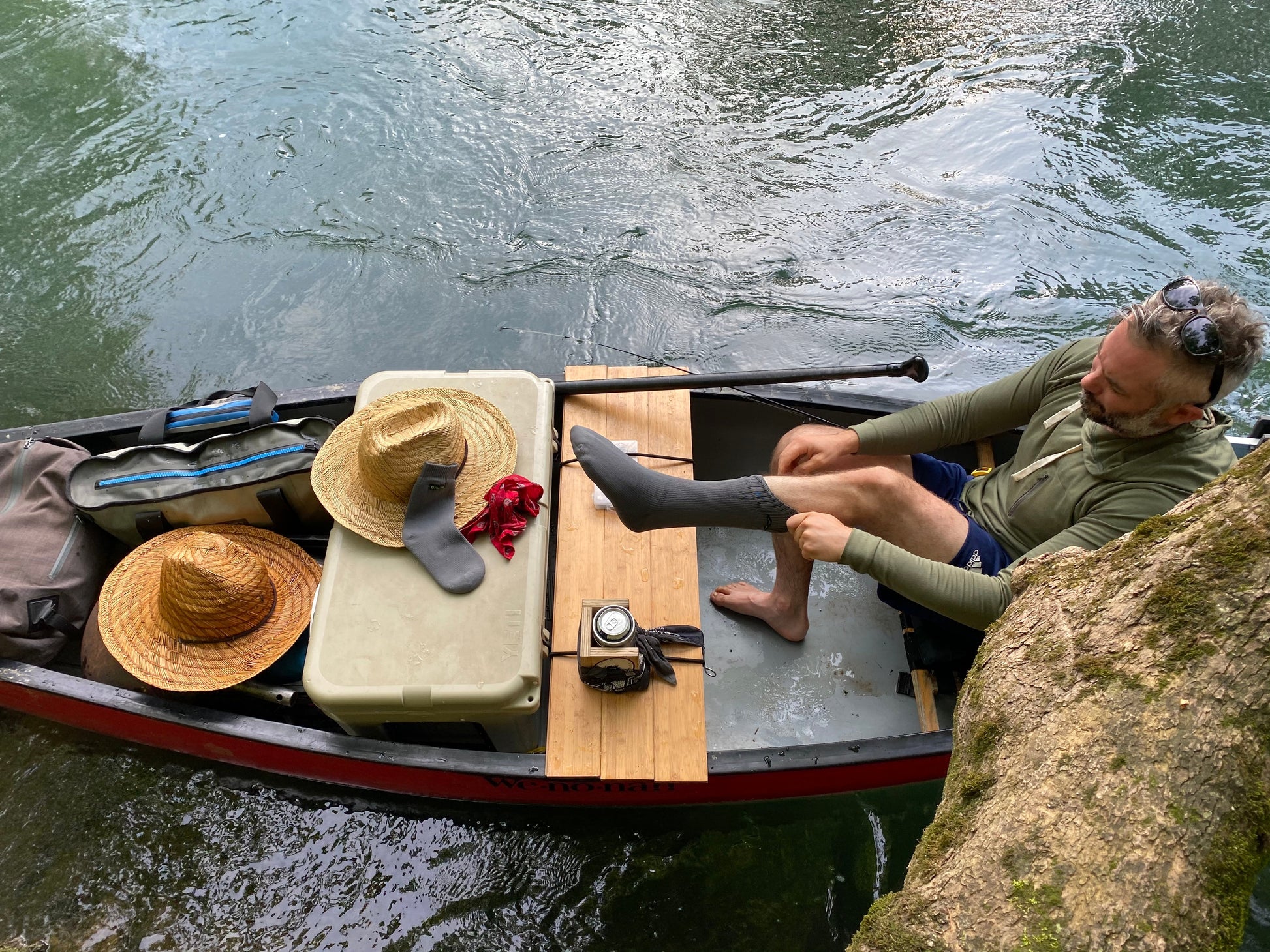 A man sits in a canoe on a river, putting on Pike Trail Waterproof Socks for blister protection. The canoe holds hats, a cooler, a camera, and other gear as sunlight reflects off the water beside him. Brand: Pike Trail.