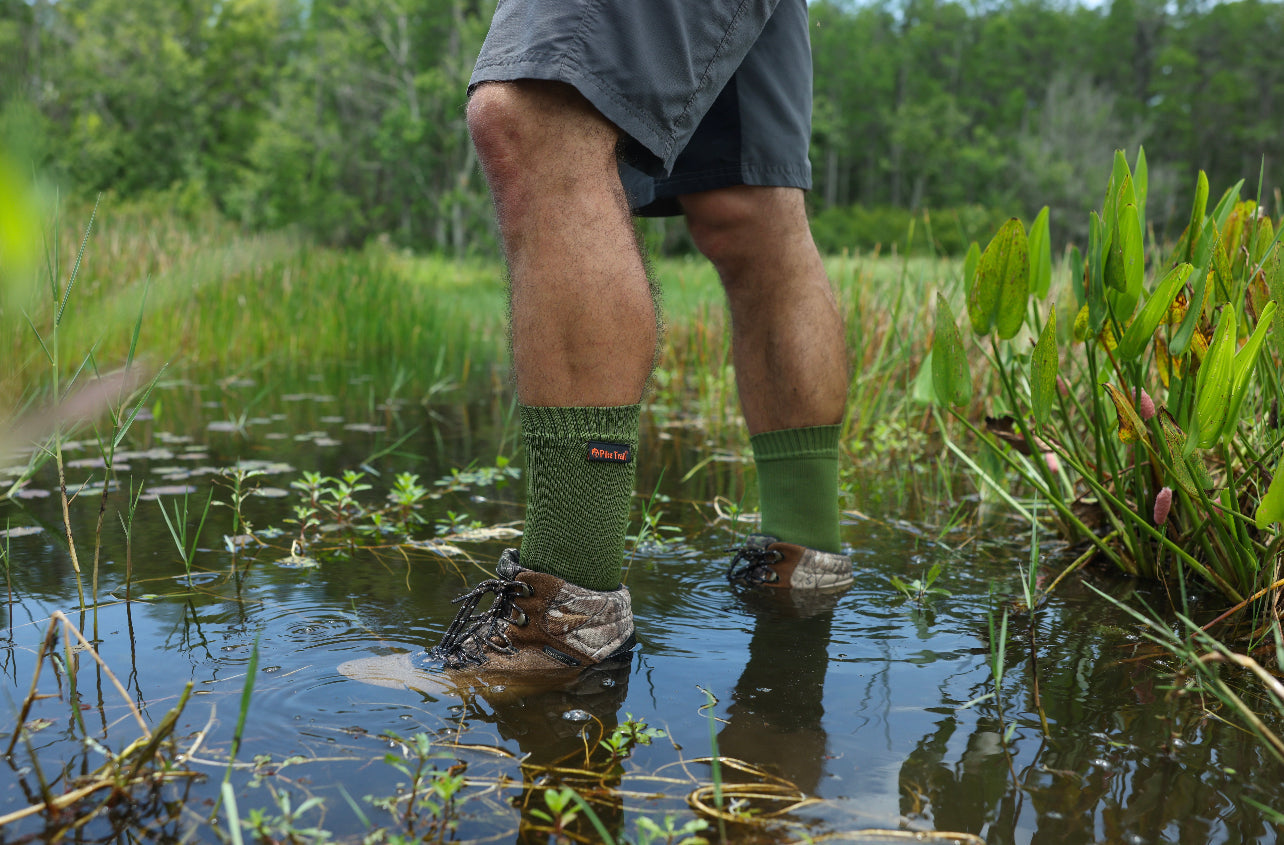 Wearing Pike Trail Waterproof Socks – Stay Dry, Stay Moving by Pike Trail, a person stands in shallow water amid grass and plants, with a forest in the background.