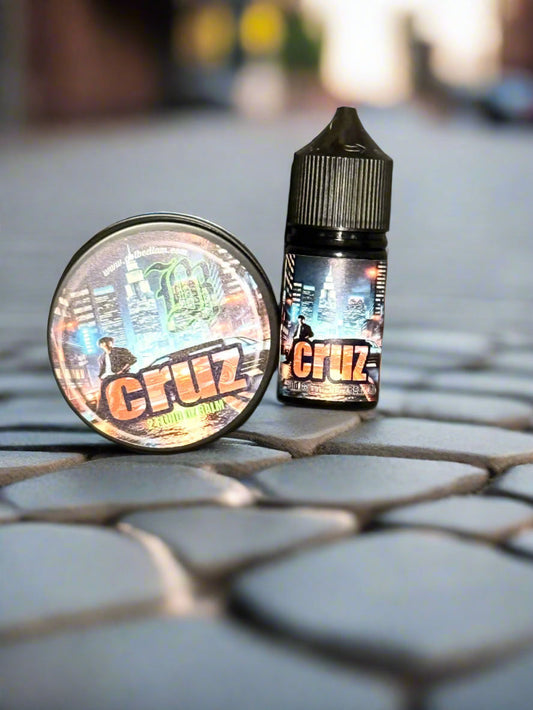 A small round tin of CRUZ Beard Balm and a dropper bottle of CRUZ Beard Oil by Bedlam Beard Company, both featuring colorful urban graphics, rest on cobblestones with a blurred cityscape in the background.