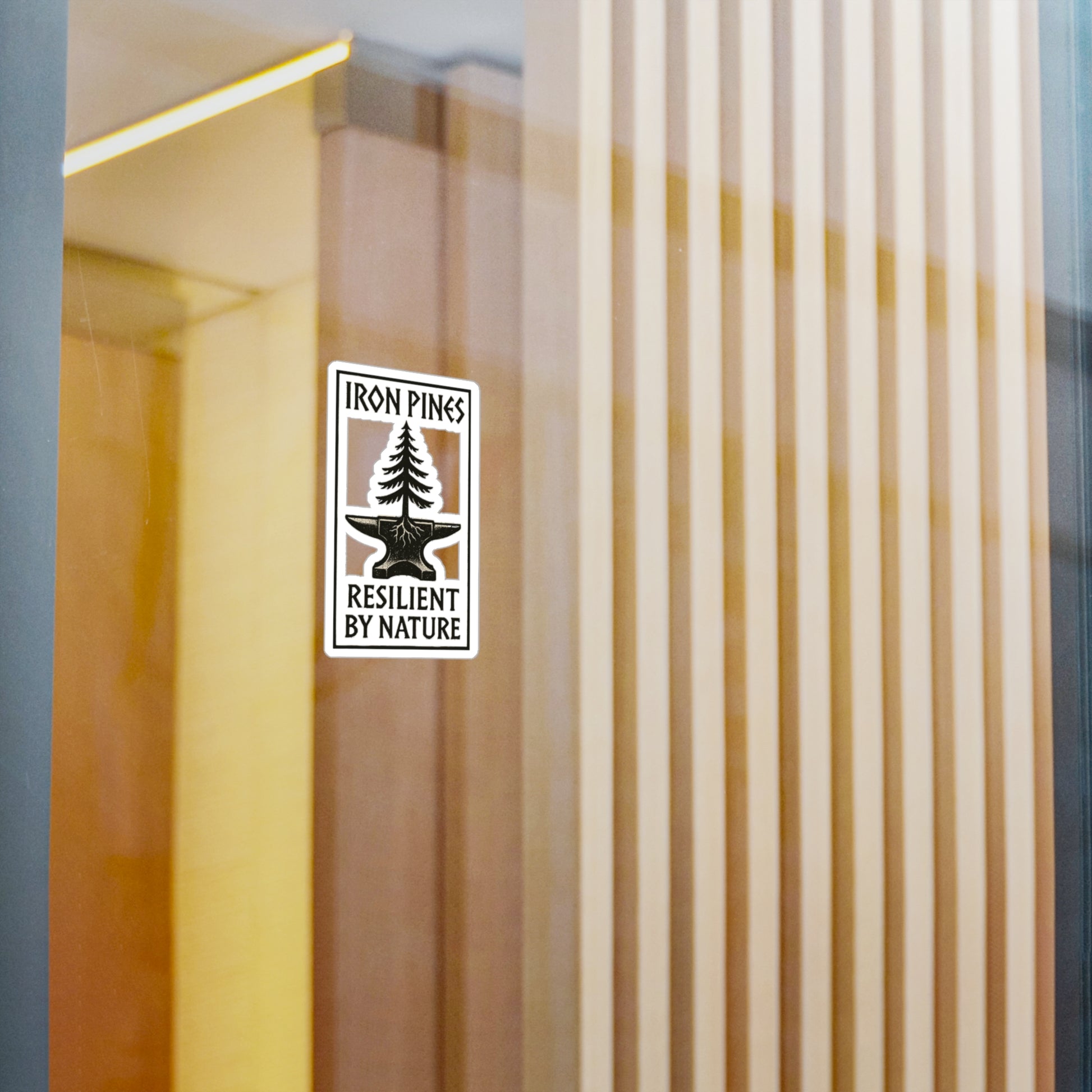 A durable vinyl Iron Pines Logo Sticker from Printify, featuring a pine tree with Iron Pines above and Resilient by Nature below, decorates a glass wall. Wooden slats and reflections in the background evoke resilience.