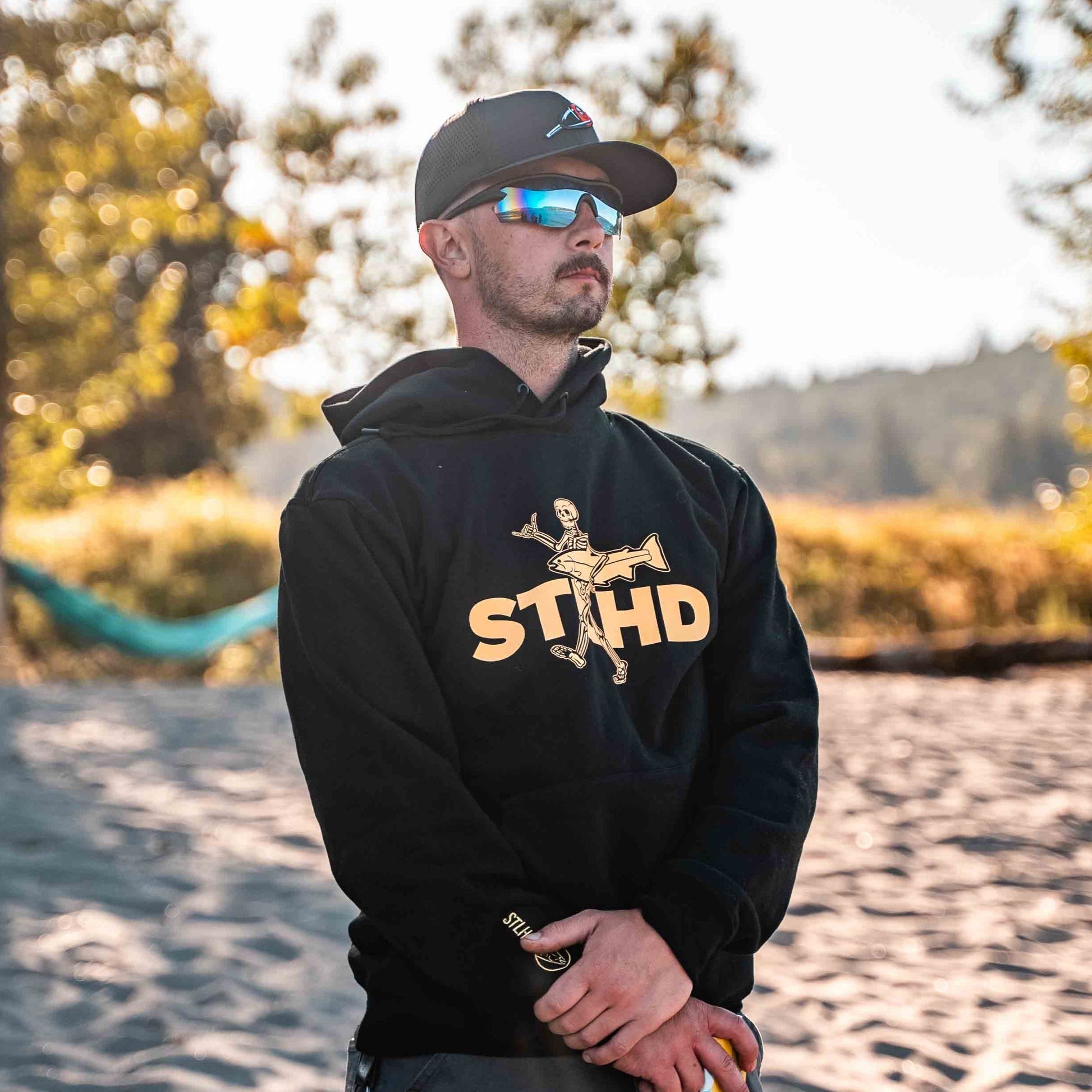 A man in sunglasses and a cap stands on sandy ground, sunlight and trees behind him, wearing the STLHD GEAR Loosey Goosey Premium Hoodie—its ultra-soft fleece offers comfort for his casual outdoor look.