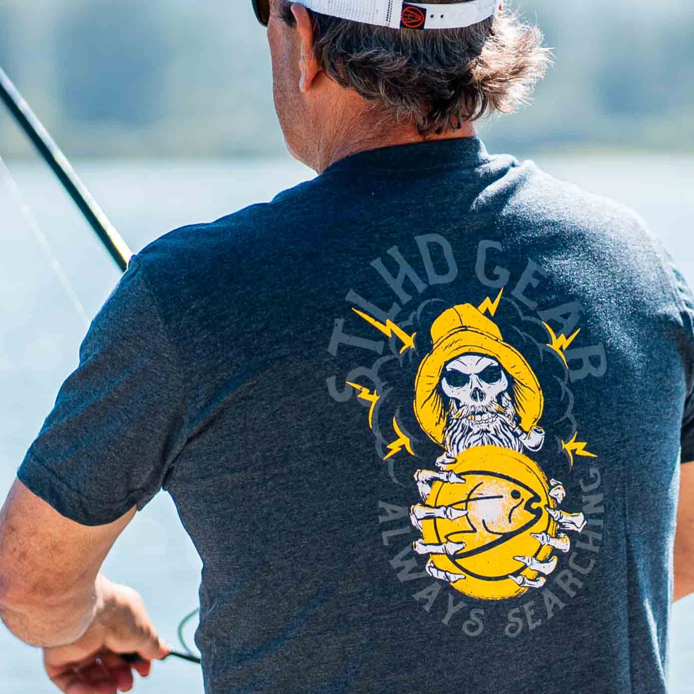 Man wearing a blue t-shirt with a graphic design on the back featuring a skull in a fisherman's hat and STLHD GEAR ALWAYS SEARCHING text, standing by a body of water.
