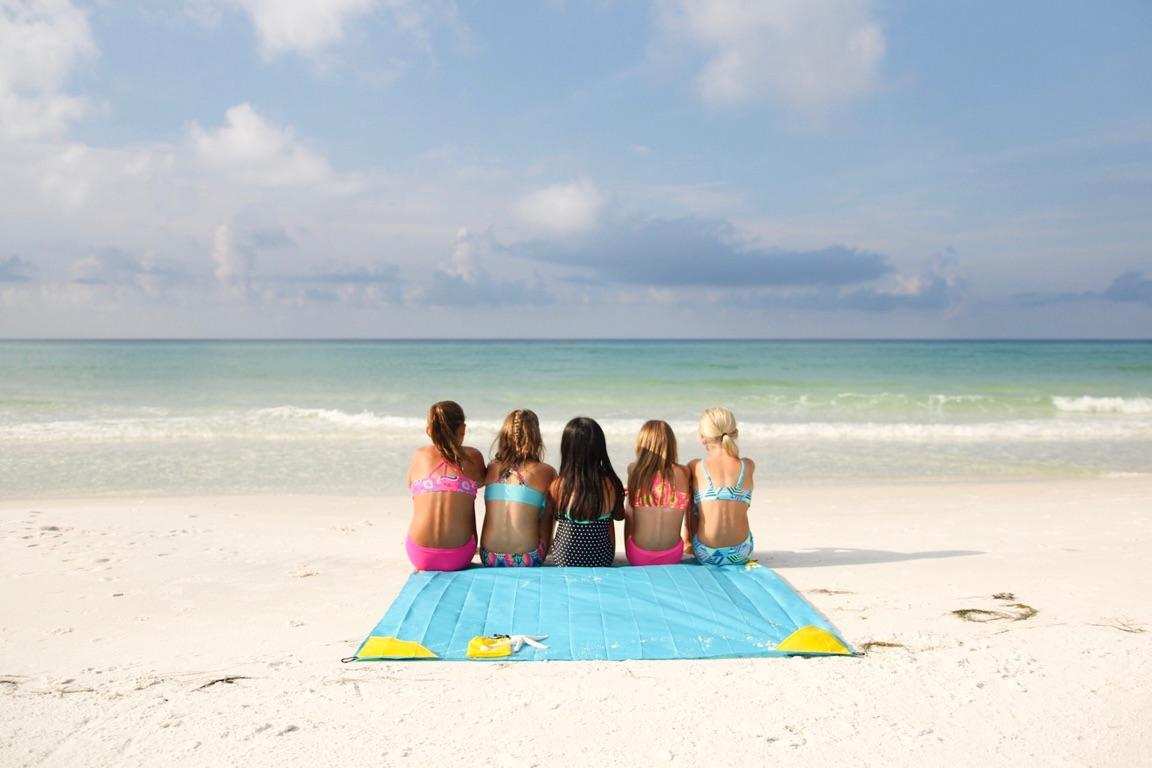 Five girls in swimsuits sit on a Pike Trail Pocket Blanket by Pike Trail, facing the ocean with their backs to the camera, on a sandy beach beneath a partly cloudy sky.