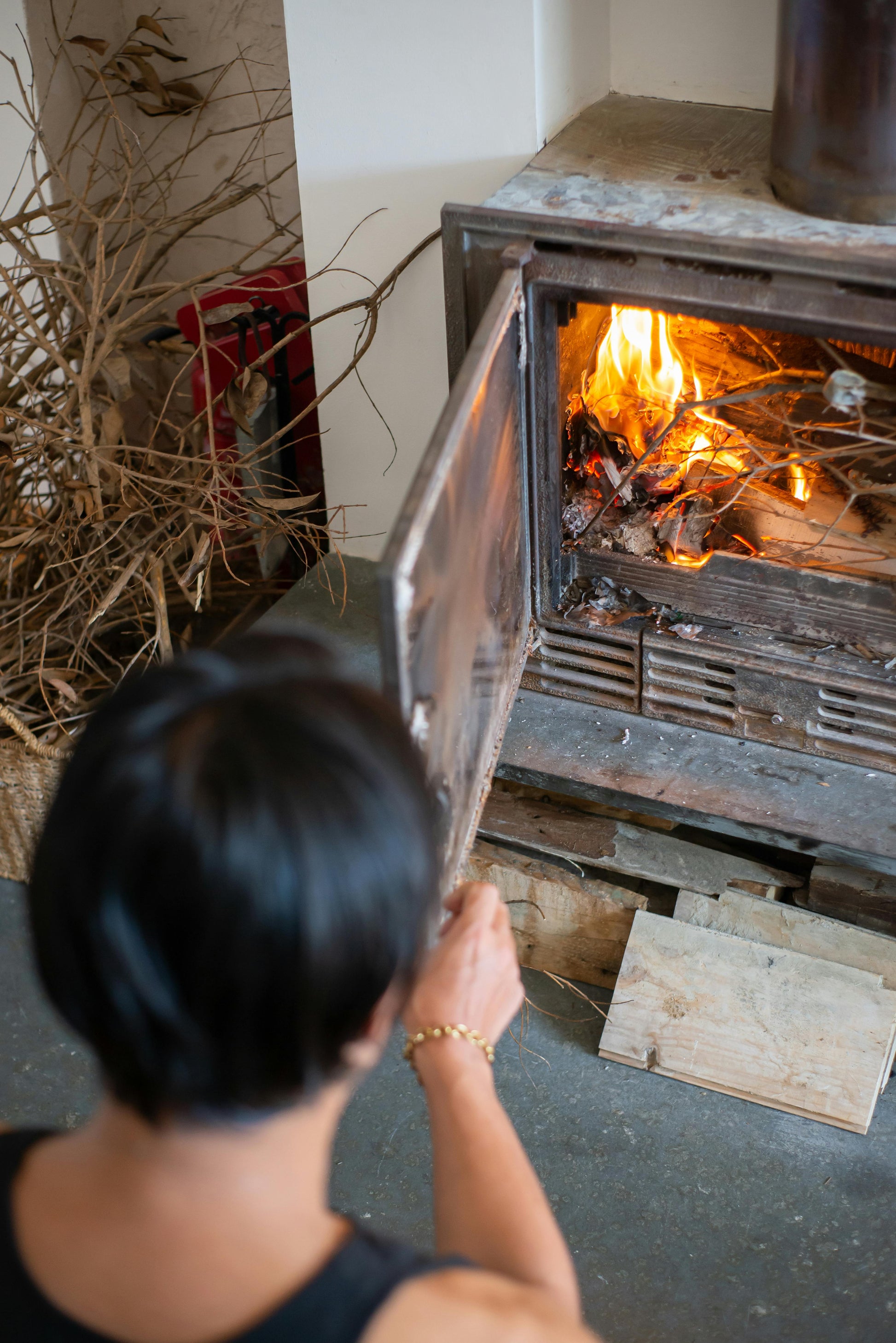 A person with short dark hair opens a wood-burning stove and adds kindling and Black Beard Fire Starters Fire Plugs - Fire Starter Tinder to a small indoor fire, while stacked branches are visible nearby.