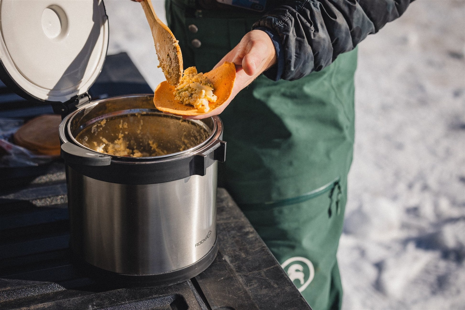 A person serves food from the RockPot Mini by RockPot onto a wooden plate outdoors using portable survival cookware, with snow in the background, wearing green pants and a black jacket.
