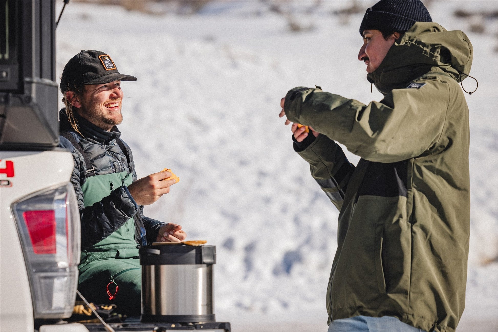 Two men in winter clothing enjoy snacks by their vehicle’s open tailgate in the snow, using the RockPot Mini by RockPot—off-grid cooking, simplified—as one sits and the other stands holding food.