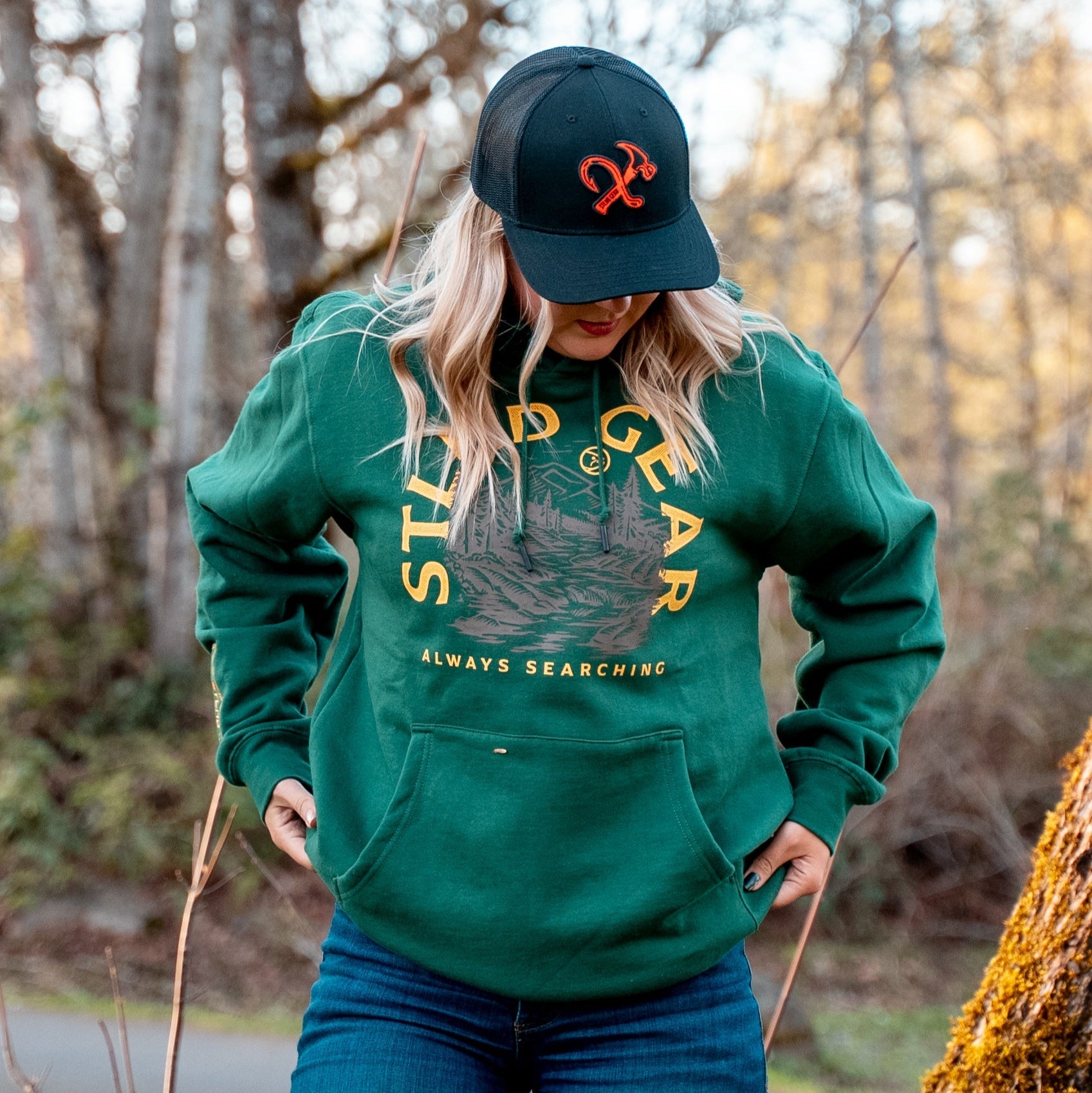 A person with long blonde hair wears an STLHD GEAR Mountain View Premium Hoodie featuring “STLHD GEAR Always Searching” and a graphic, paired with a black cap, while standing outdoors in a wooded area, looking down.