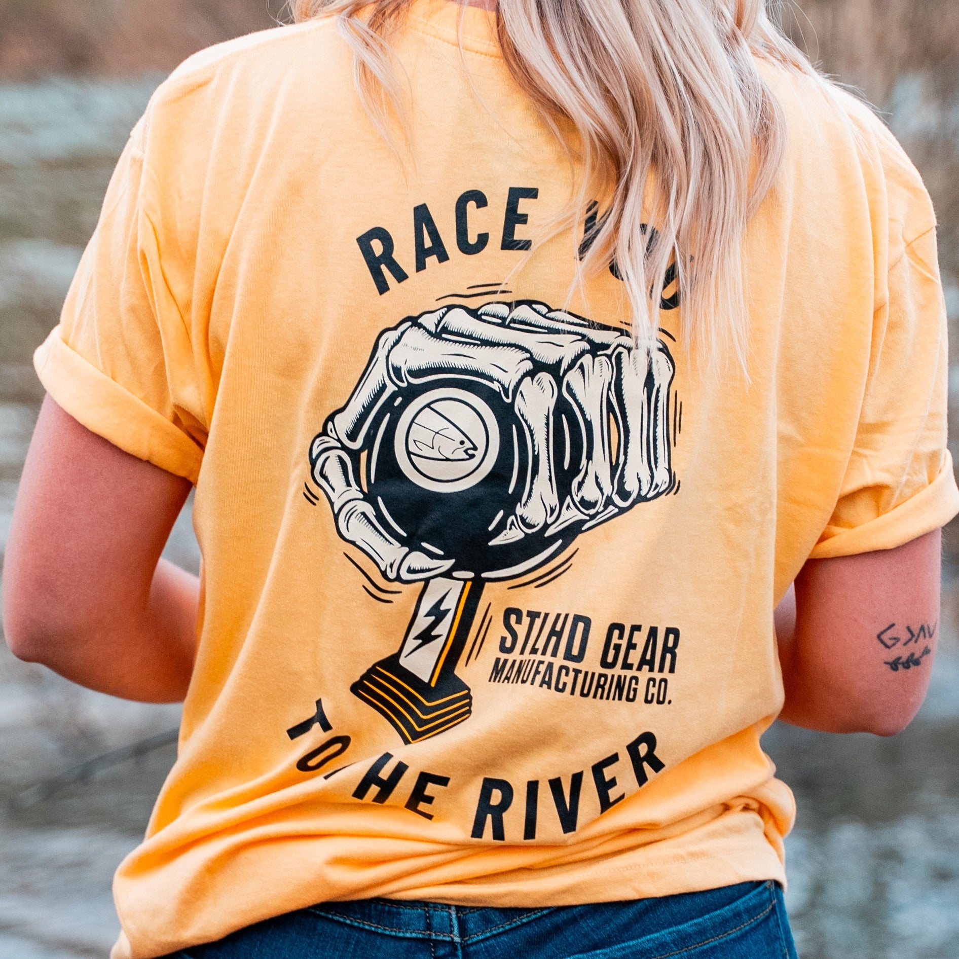 Wearing the yellow STLHD Race Ya T-Shirt from STLHD GEAR, featuring a piston illustration and “RACE TO THE RIVER” on the back, someone enjoys the riverside view in this comfortable, breathable shirt.