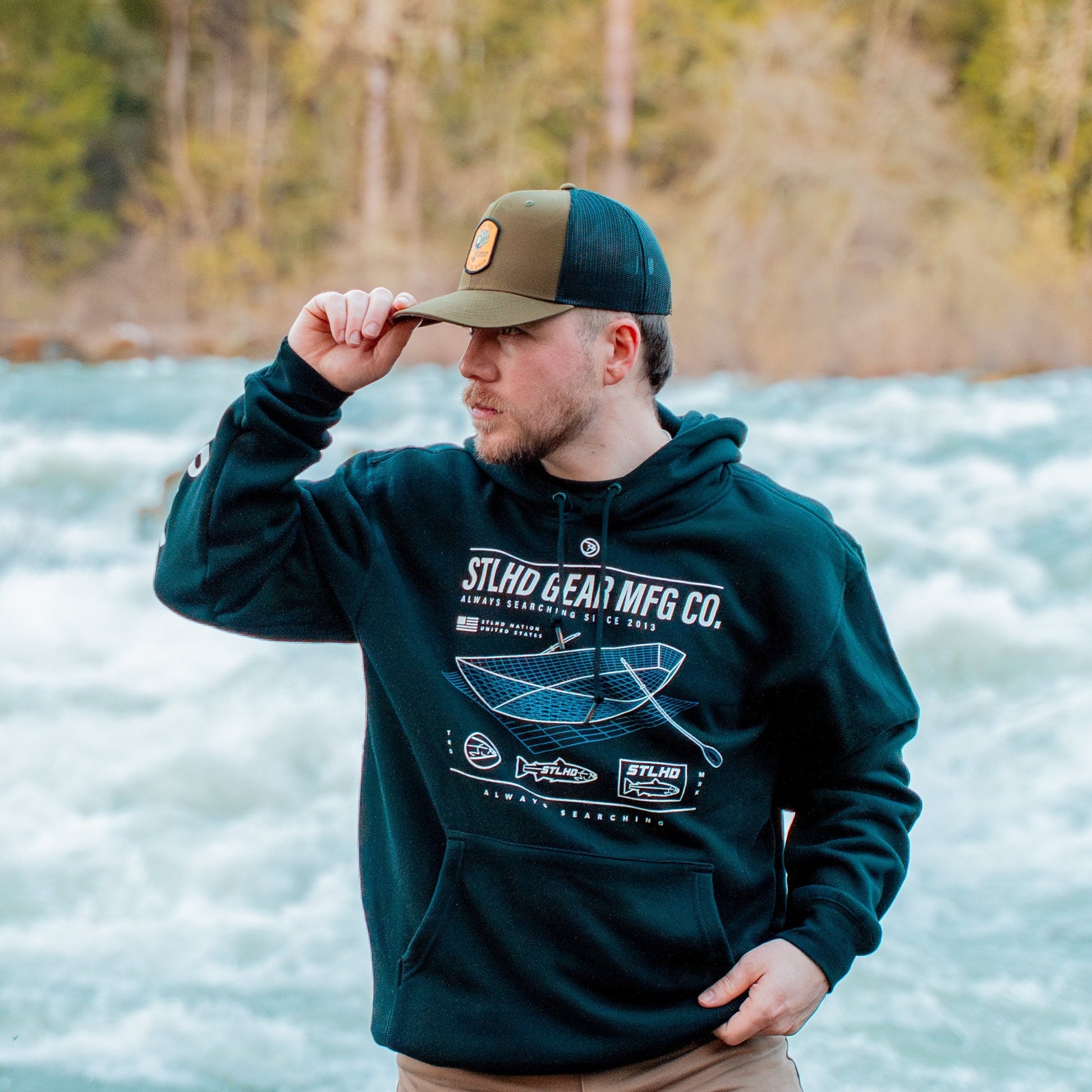 A man in an STLHD GEAR STLHD Tech Spec Premium Hoodie and green cap stands by a fast-flowing river, holding his cap’s brim. Blurred trees and rocks appear in the background.