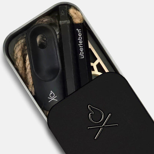 The Überleben FIREKIT – All‑in‑One Fire Starter Kit features a compact black sliding lid, with the Überleben logo, and includes fire-starting tools plus natural tinder, all neatly organized for your next survival adventure.
