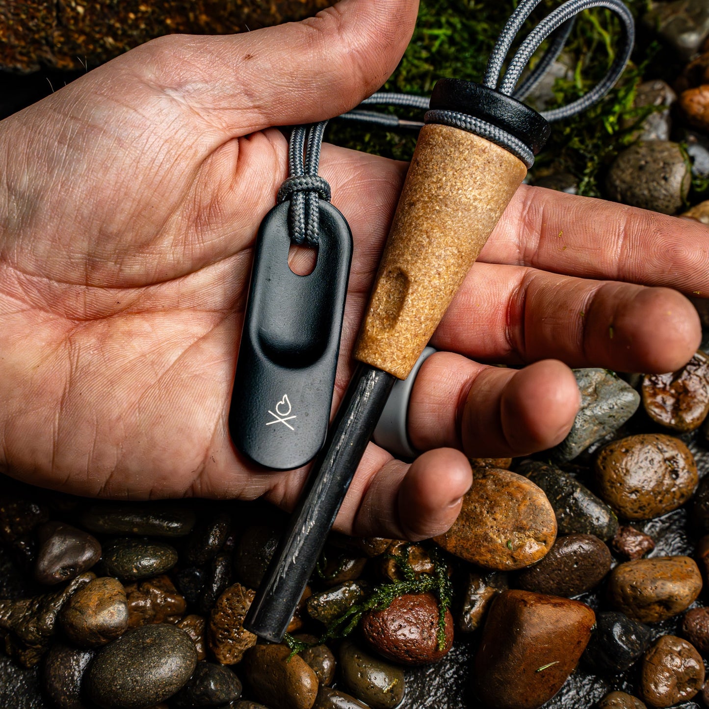 A hand holds the Überleben Zünden PRO – Modern Flint & Steel Fire Starter by Überleben, featuring a wood-composite handle and black striker on gray cords, above wet river stones.
