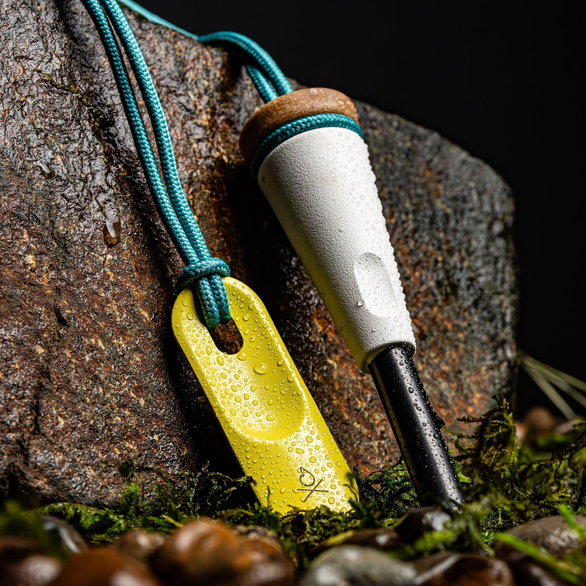 The Überleben Zünden PRO Modern Flint & Steel Fire Starter by Überleben, featuring a white handle, cork top, and black ferro rod on a blue cord with yellow striker, rests on mossy rocks with water droplets—perfect for outdoor adventure.