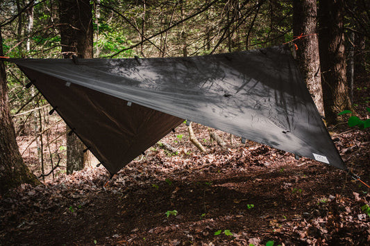 The TITAN Survival TACAMO® Bushcraft Reflective Shelter Tarp is set up between trees in a forest, creating shade on the leafy forest floor and secured with ropes tied to tree trunks.