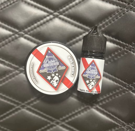 A tin of beard balm and a small bottle of beard oil labeled Bedlam Beard Company CABLE - Warm & Spicy Blend rest on quilted black leather. The labels have a red stripe, blue emblem, and promise a bold masculine scent.