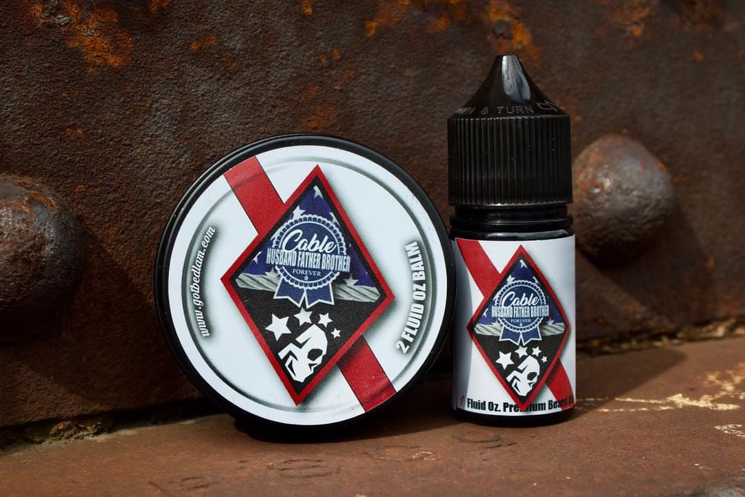 A bottle of CABLE - Warm & Spicy Blend beard oil and balm from Bedlam Beard Company, with patriotic labels (stars, stripes, bearded face logo), set on rusty metal. These products deliver a bold masculine scent.