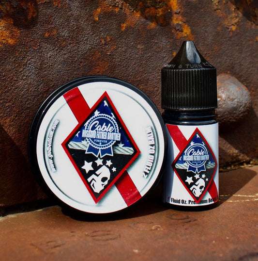 A round tin of beard balm and a small bottle of beard oil from Bedlam Beard Company, both labeled “CABLE - Warm & Spicy Blend” with blue, red, and white patriotic designs featuring stars and a skull, rest on a rusty metal surface.