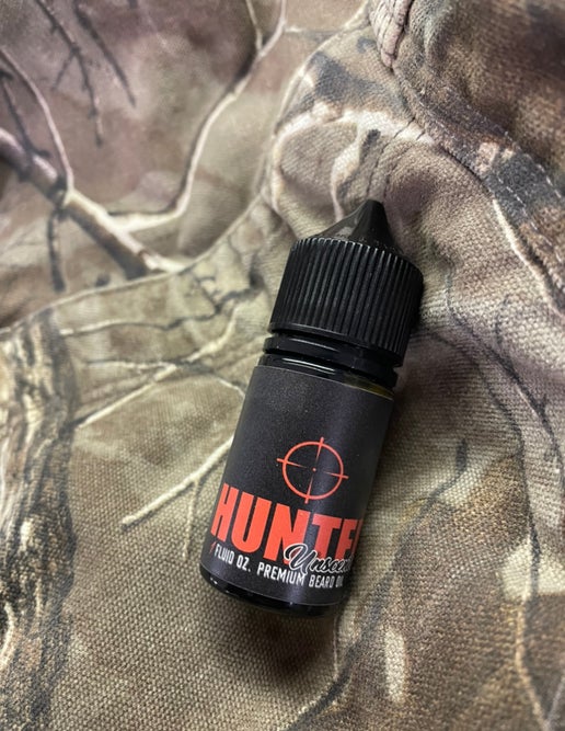 A small black bottle labeled Hunter – Unscented Beard Oil from Bedlam Beard Company, with a red crosshair, rests on camo fabric—premium field-grade oil ideal for hunters seeking quality grooming in the field.
