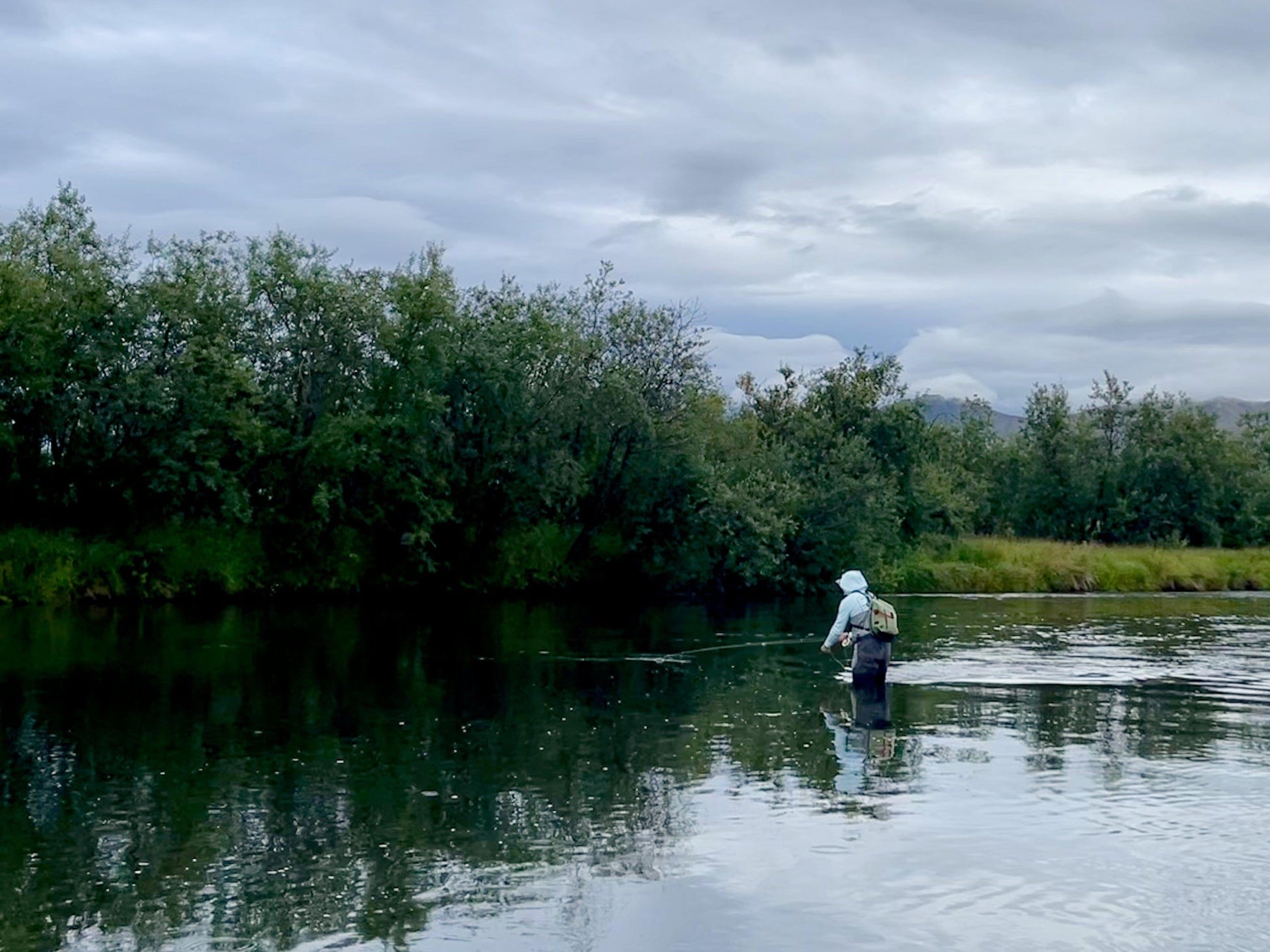 An angler wearing waders and a hooded jacket fly fishes by a tree-lined riverbank under cloudy skies, carrying the Jackson Hole Fly Company JHFLYCO 20L Waterproof Sling Pack—streamlined for the mission—over their shoulder.