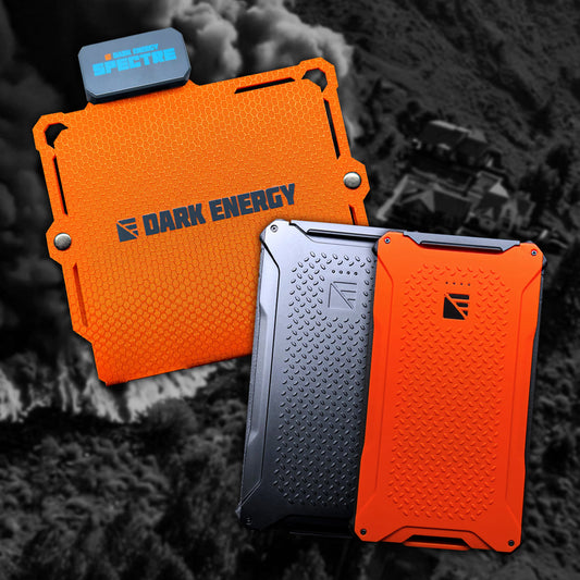 Three rugged power banks from the Entropy Survival Off Grid Survival Power Bundle—two orange, one black—are displayed on a smoky grayscale background with a destroyed building.