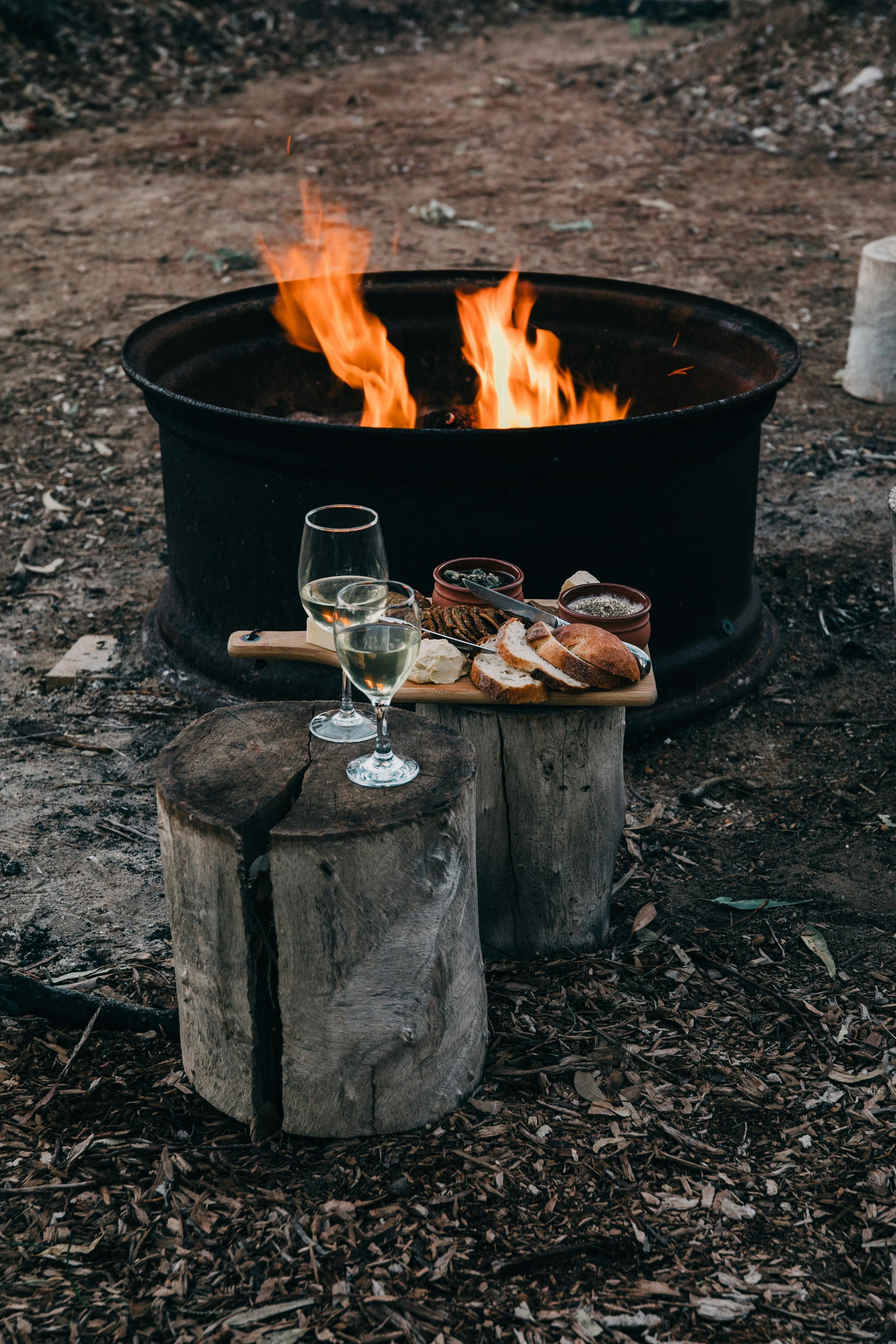 Two glasses of white wine and a wooden platter with bread and dips rest on tree stumps by a crackling fire pit, its flames fueled by Fire Plugs - Fire Starter Tinder from Black Beard Fire Starters, outdoors on dirt.
