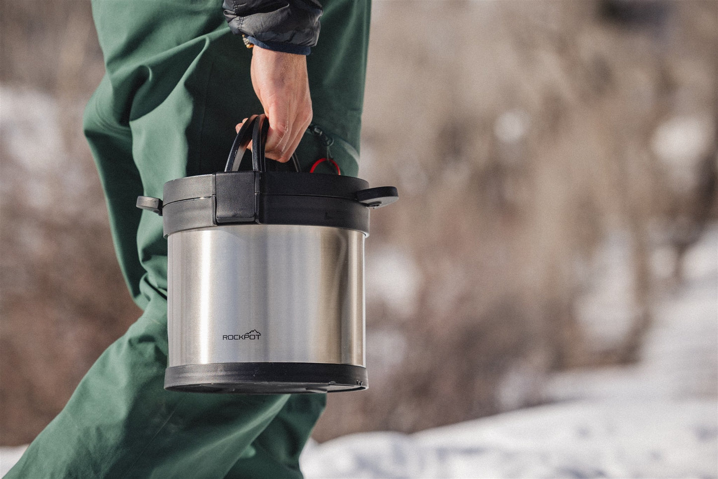 A person in green pants carries a RockPot Mini by RockPot, featuring black handles—perfect for off-grid cooking—while outdoors in a snowy setting.