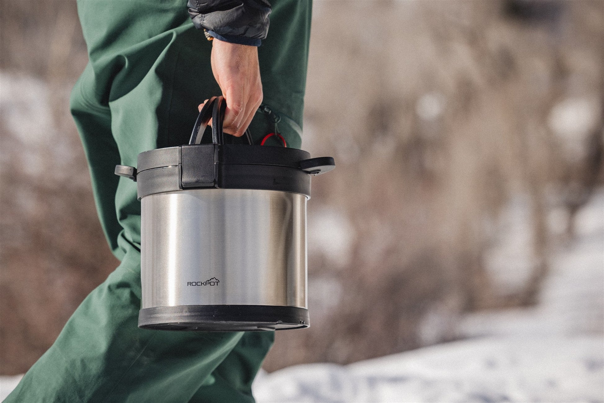 A person in green pants carries a RockPot Mini by RockPot, featuring black handles—perfect for off-grid cooking—while outdoors in a snowy setting.