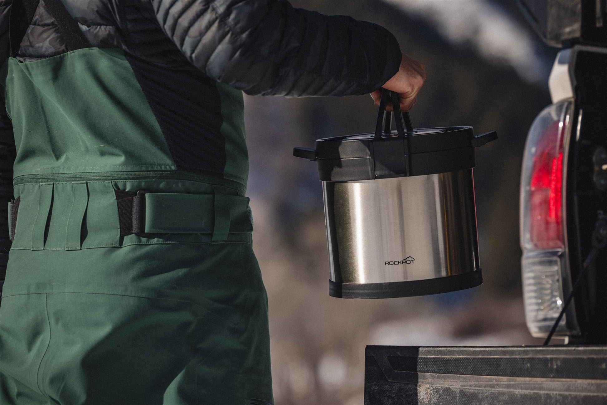 Wearing green overalls and a black jacket, a person lifts the RockPot Mini Bundle – All-In Survival Cooking, No Plug Required by RockPot from a pickup truck outdoors, with mountains and snow in the background.